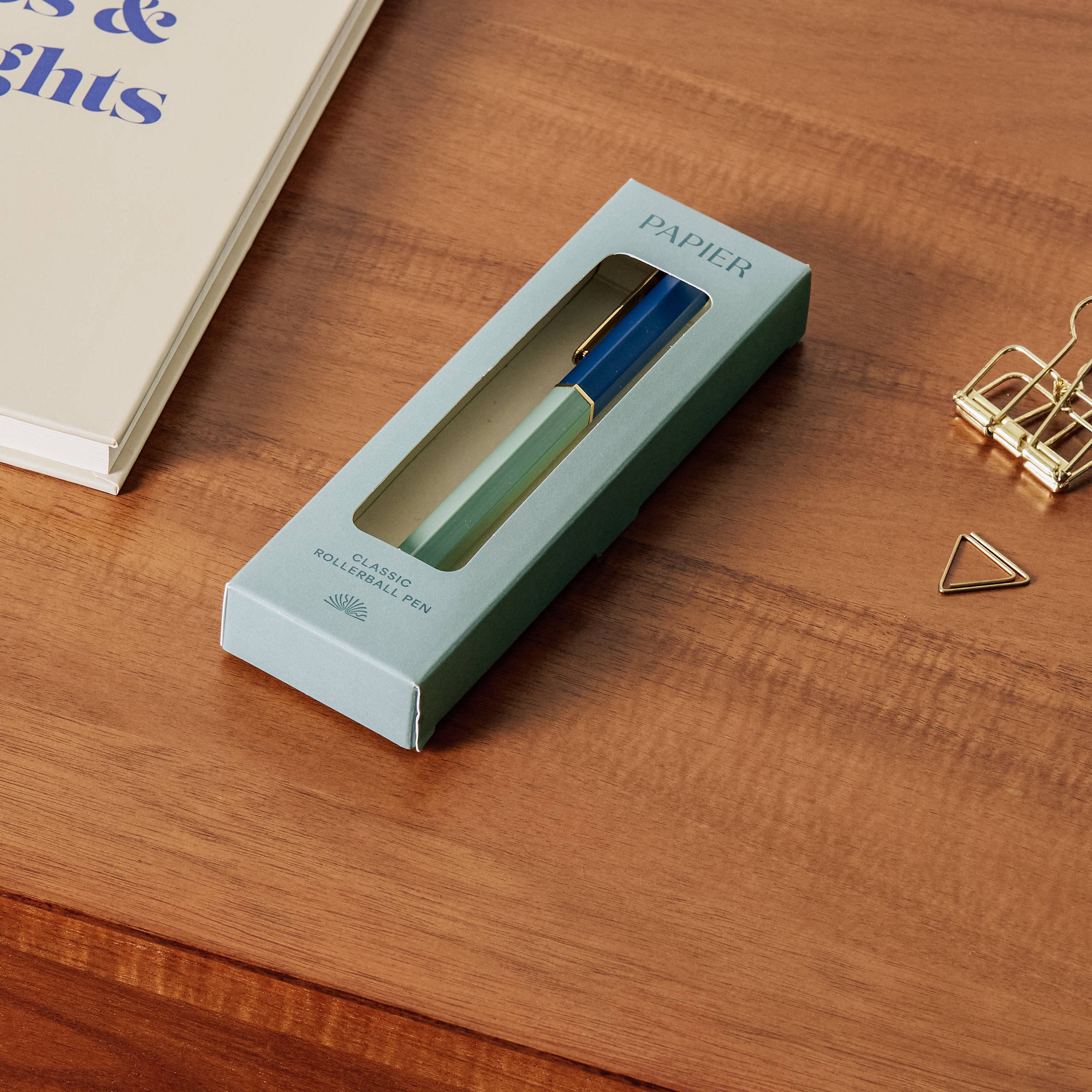 Green pen in a box on a wooden surface with a book and gold clip in the background