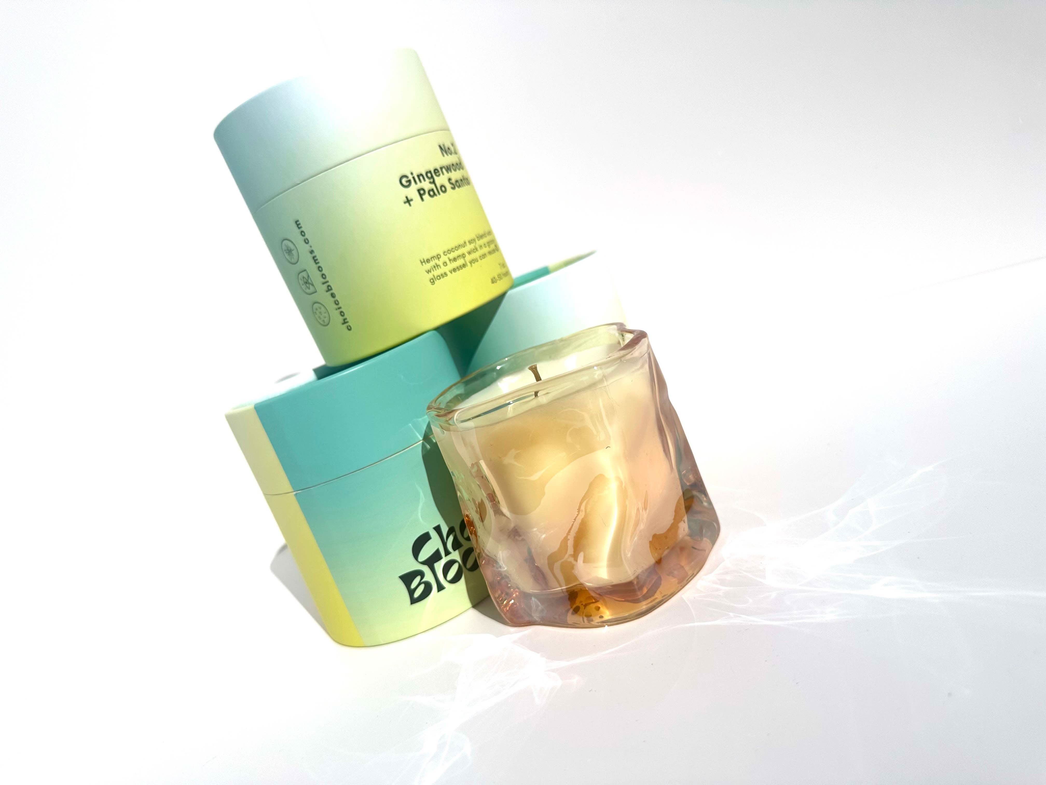 One candle in tinted pink wavy glass container next to three Aqua and green candle packaging containers 