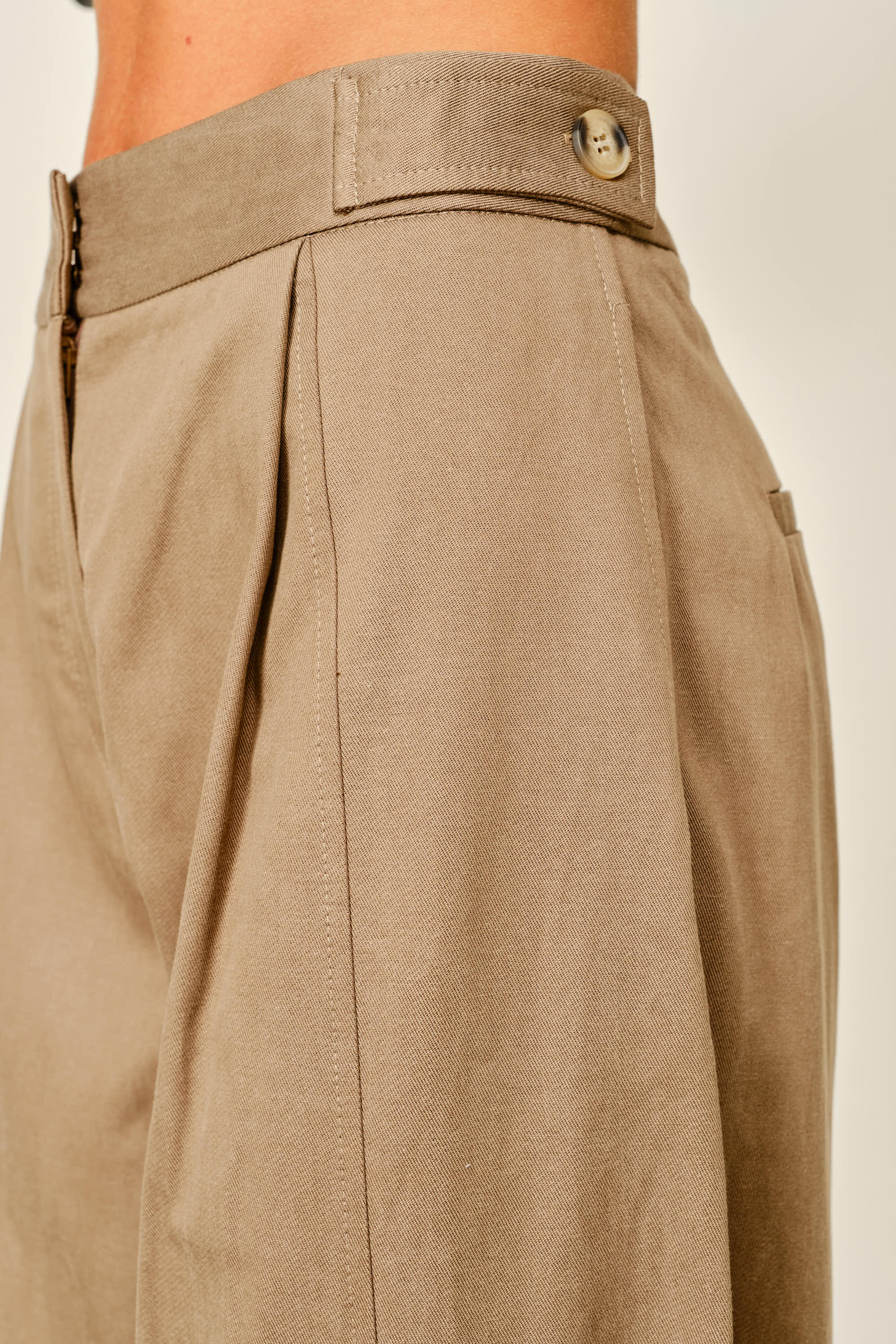 Close-up of a model wearing high-waisted taupe trousers, showing the waistband with a button closure and pleated front detail.