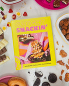 Bright yellow and pink cover of cookbook “caught snackin” featuring chicken and waffles on the cover of the book surrounded by assorted snack 