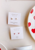 Two sets of earrings, tiny heart-shaped opals set in a gold three-prong stud, one set pink, one set white and displayed on earring card