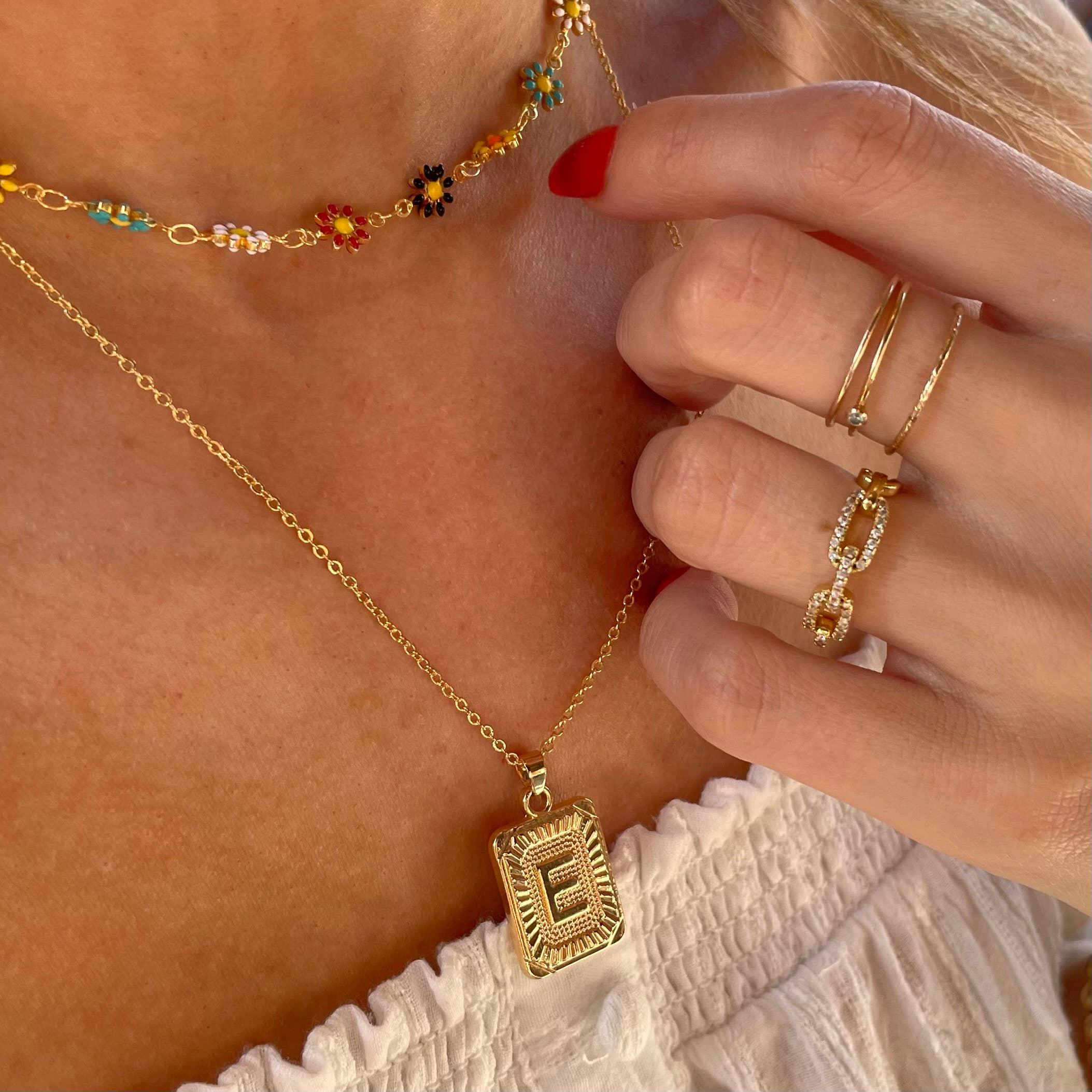 Woman wearing a letter E tarnish free gold initial charm necklace with a colorful flower necklace while wearing gold rings. 