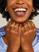 Smiling woman wearing blue shirt with hands under her chin wearing gold letter rings in the words BE  BOLD 