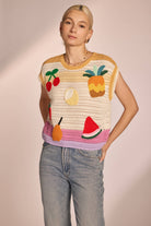 Woman wearing sleeveless, ivory, colorful, crochet, top with cherries, lemon, strawberry, watermelon, pear, and pineapple with pink, purple, and yellow horizontal stripes 