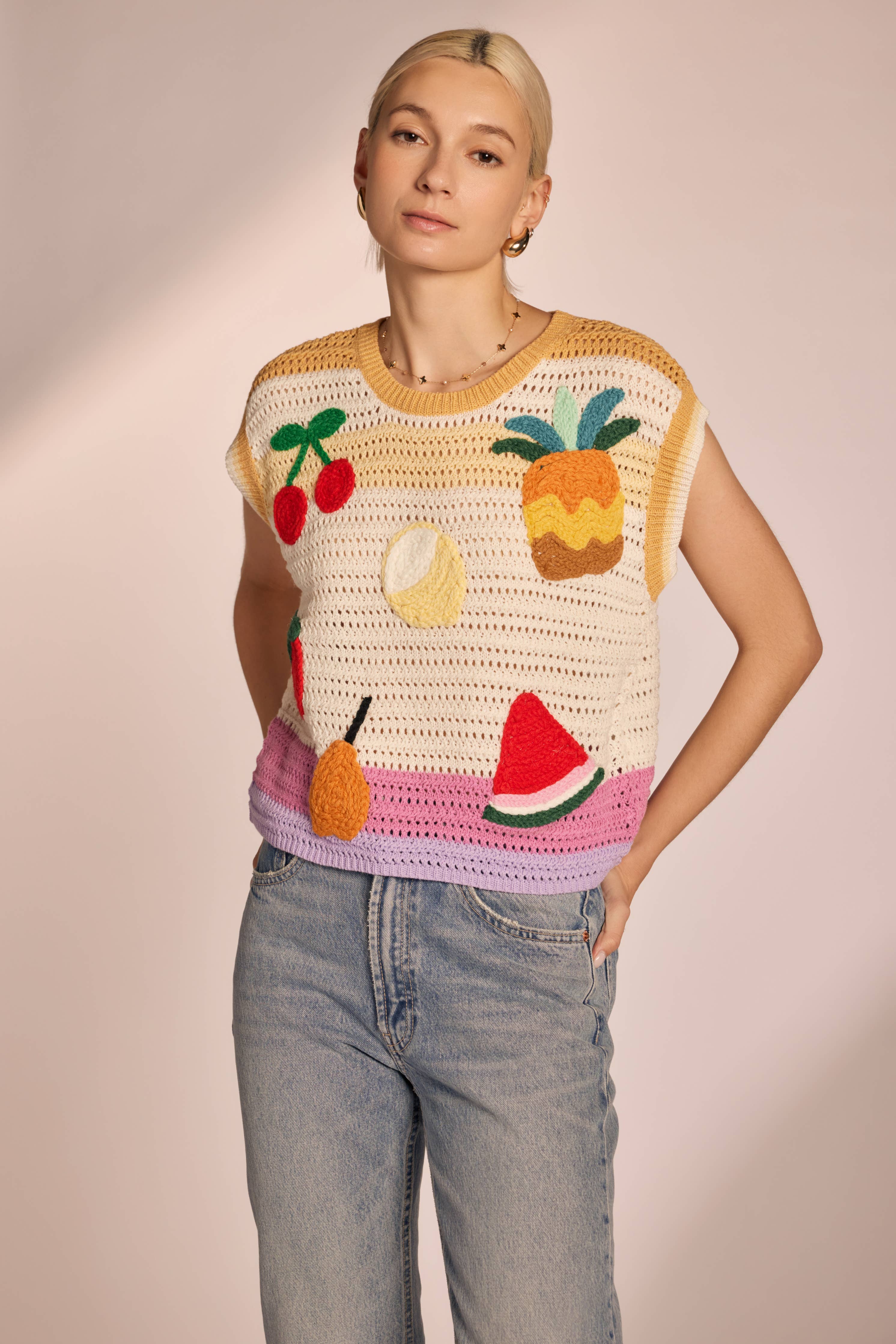 Woman wearing sleeveless, ivory, colorful, crochet, top with cherries, lemon, strawberry, watermelon, pear, and pineapple with pink, purple, and yellow horizontal stripes 