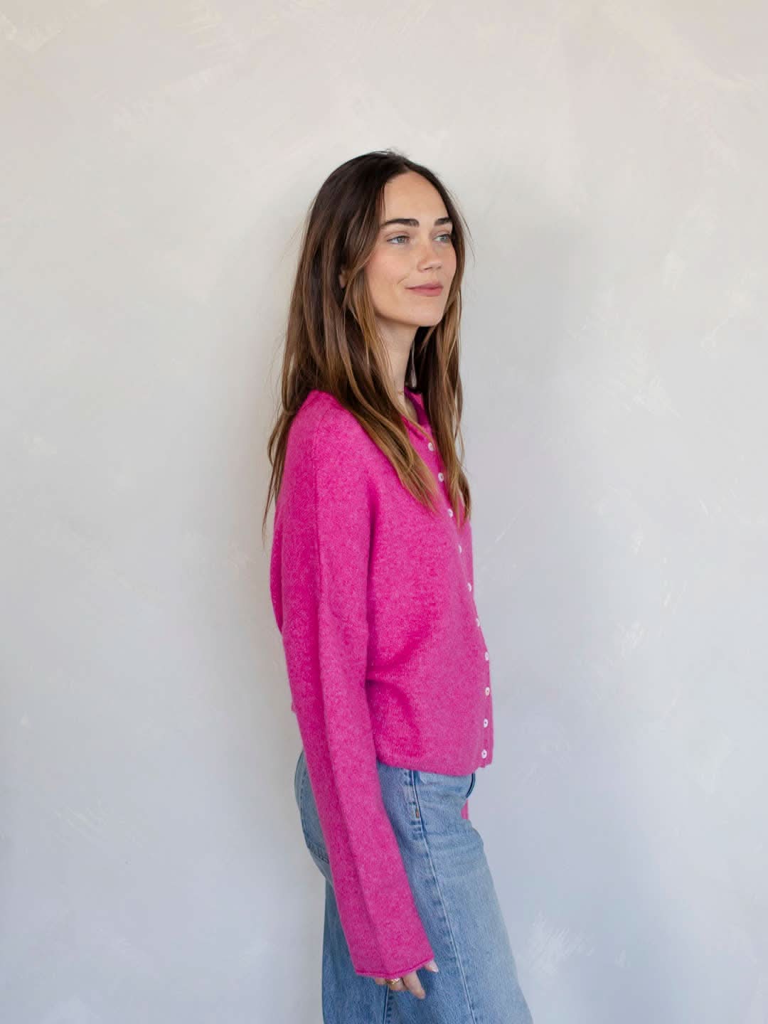 Woman smiling standing against white background wearing Fuschia pinkish button down cardigan with white buttons and blue jeans