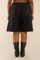 Woman wearing black vertical pin striped knee length shorts and black boots behind a blank background. 