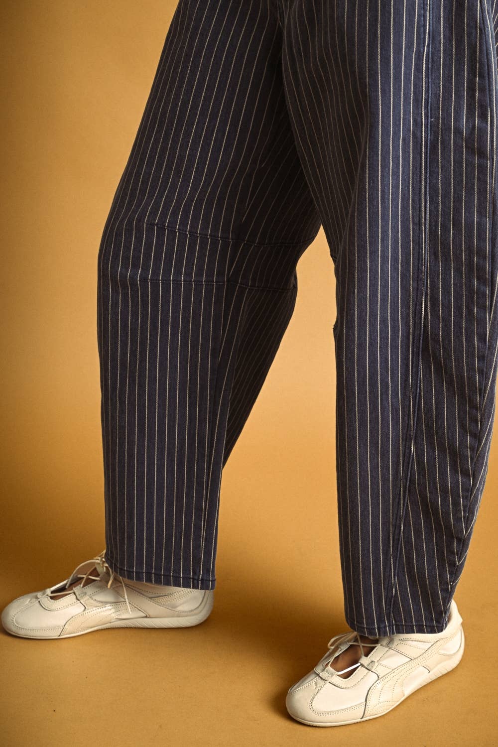 A person wearing loose navy blue pinstriped trousers and white sneakers, standing against a warm tan studio background.
