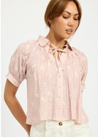 Model standing against white background wearing a pink blouse with tulip embroidery and buttons in front and tie collar. 