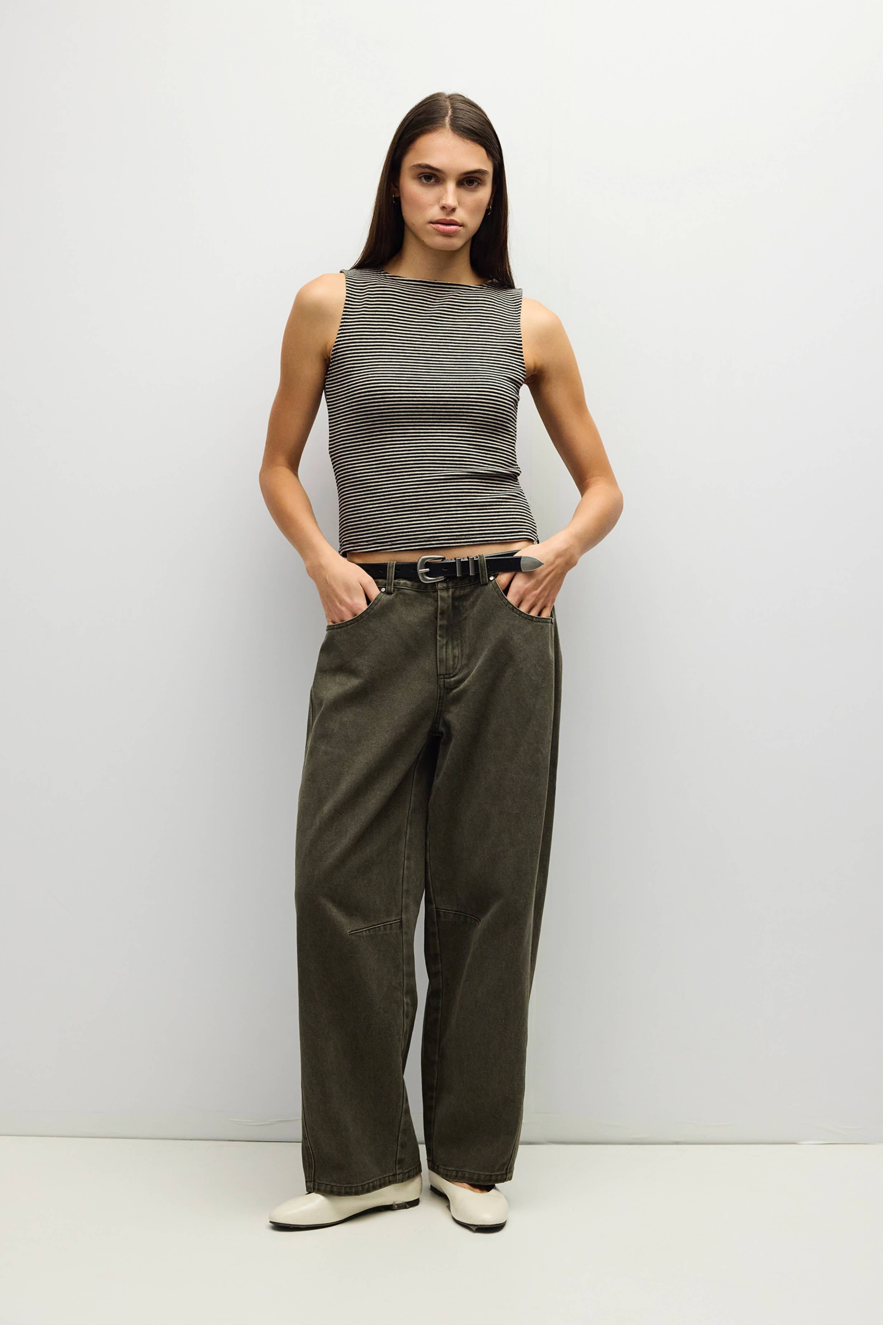 A woman stands against a plain light gray studio wall wearing a sleeveless ribbed gray top, dark olive wide-leg pants, and white shoes.