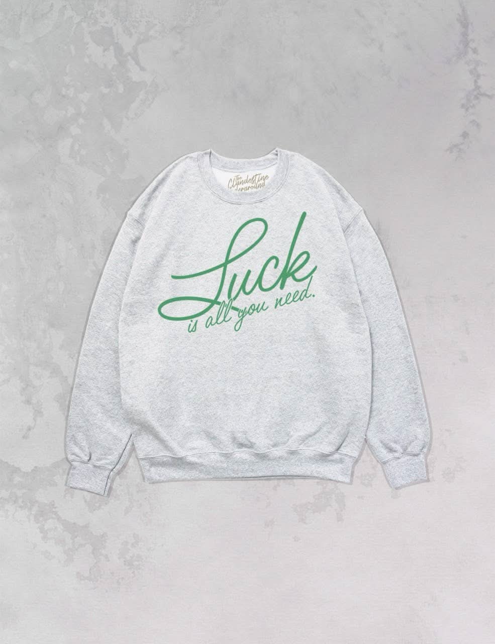 “luck is all you need” written in green cursive on grey oversized pullover sweatshirt laying in grey marbled background 