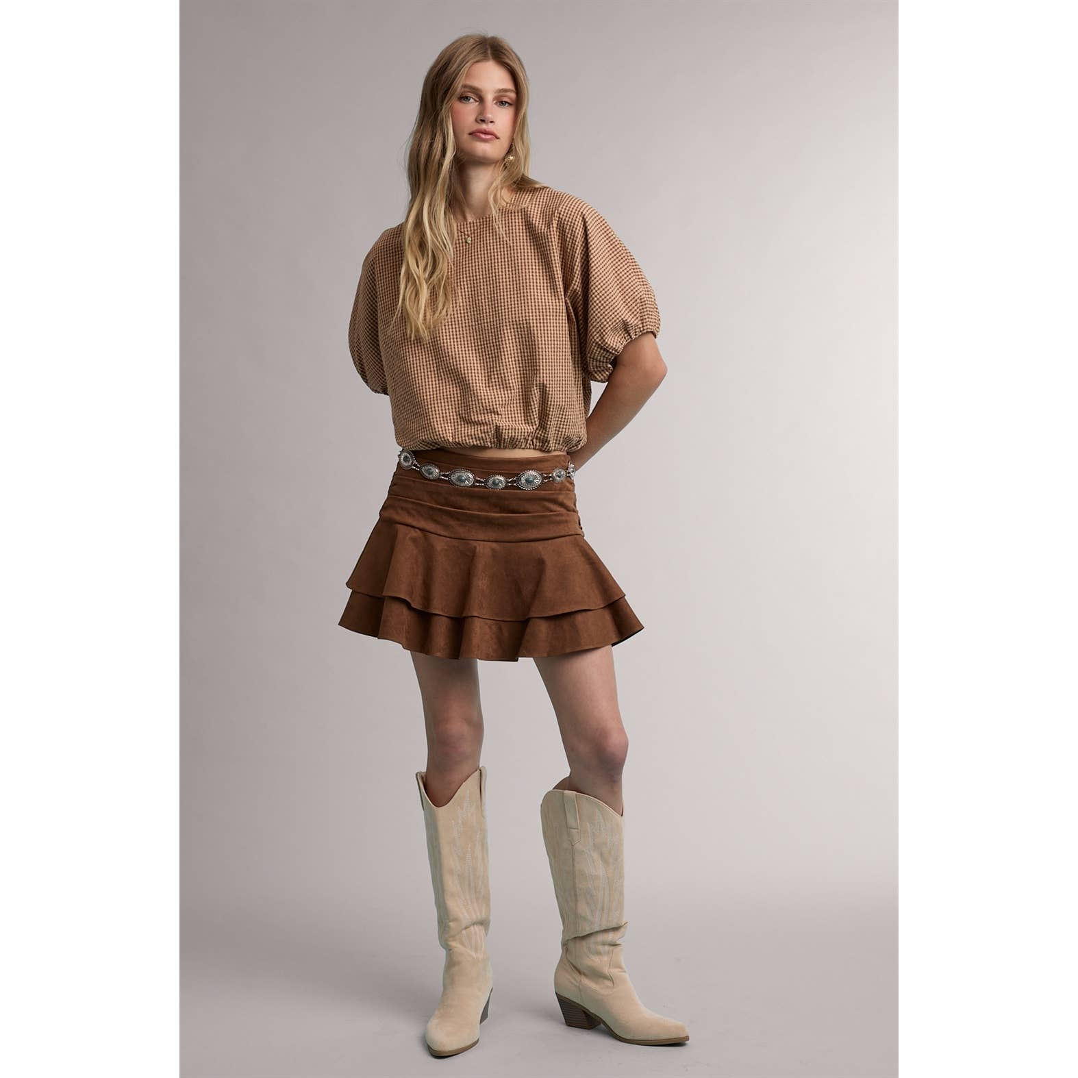 A woman modeling a brown textured short-sleeve blouse with puffed sleeves, a brown tiered ruffle mini skirt with a silver-toned patterned belt, and beige cowboy boots, set against a plain light gray studio background.