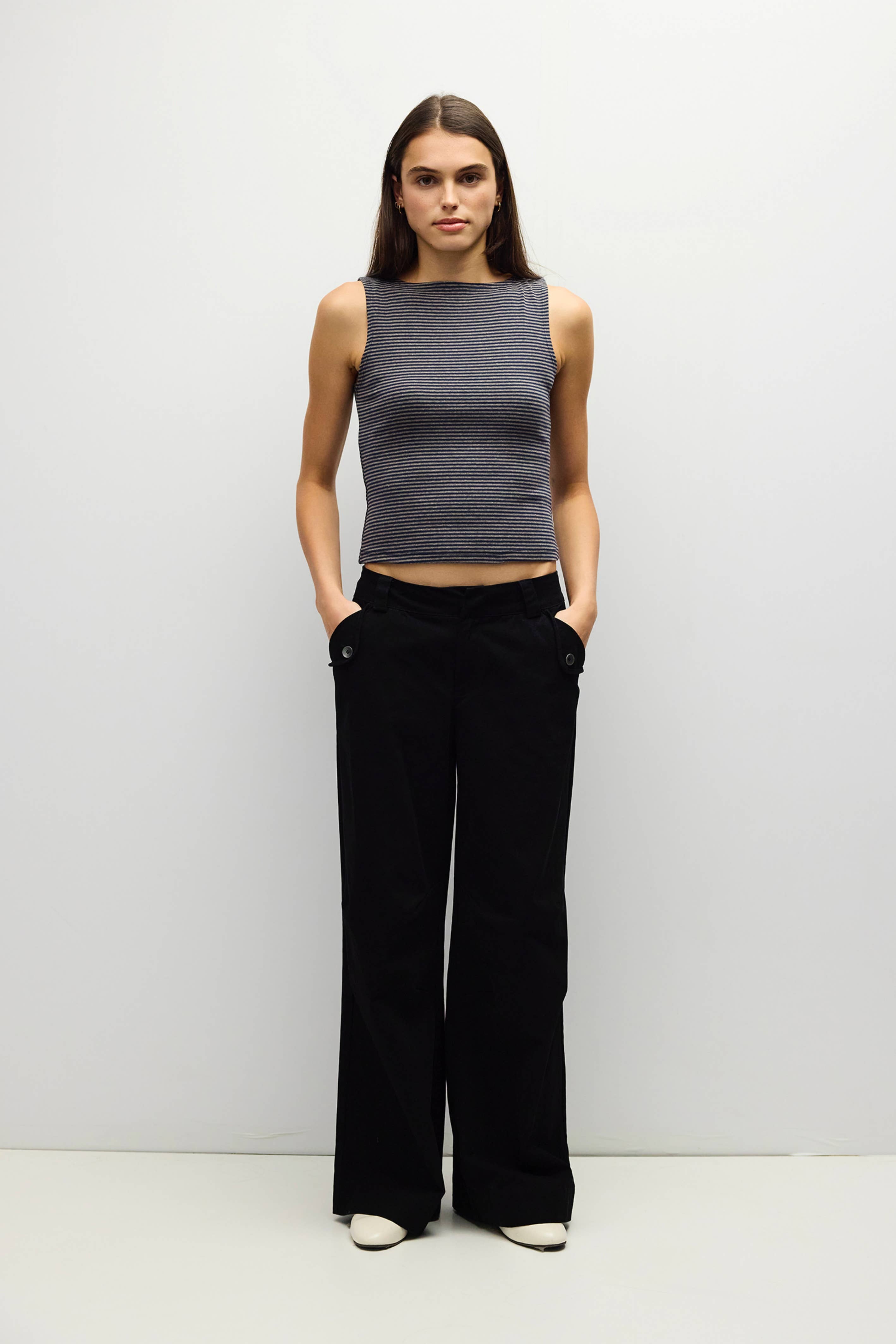 A woman stands against a plain light gray studio background wearing a sleeveless dark gray ribbed knit top and black wide-leg pants with white shoes.