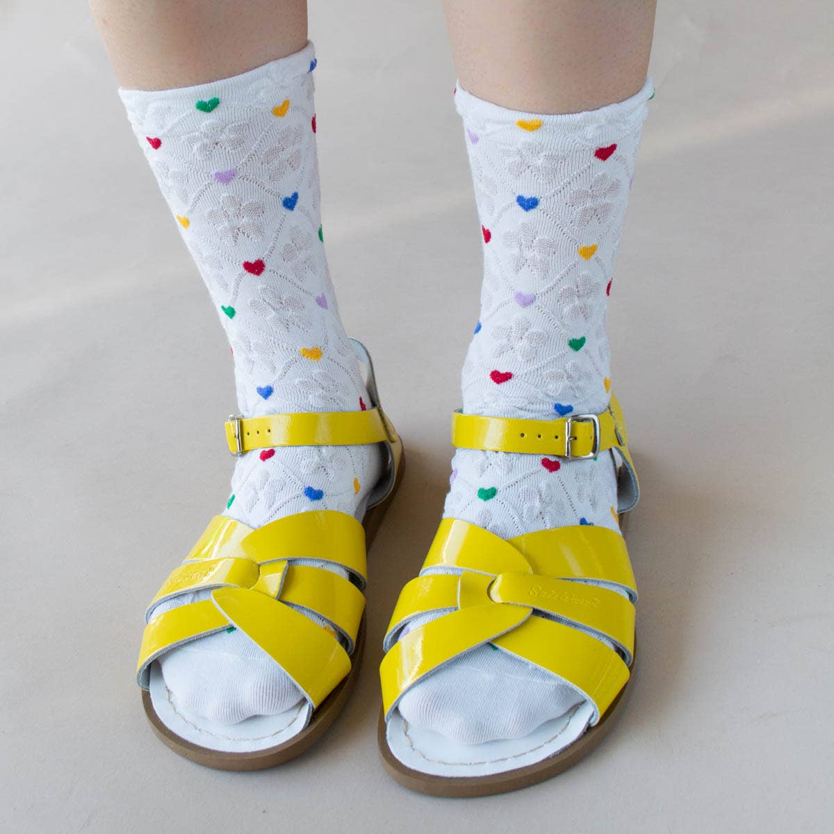 model wearing white socks with red, lilac, yellow, green, and blue hearts and wearing yellow sandals 