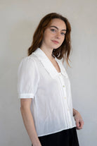 Woman standing against white background wearing black shorts and white collared button down blouse with extended sleeves and vertical line details 