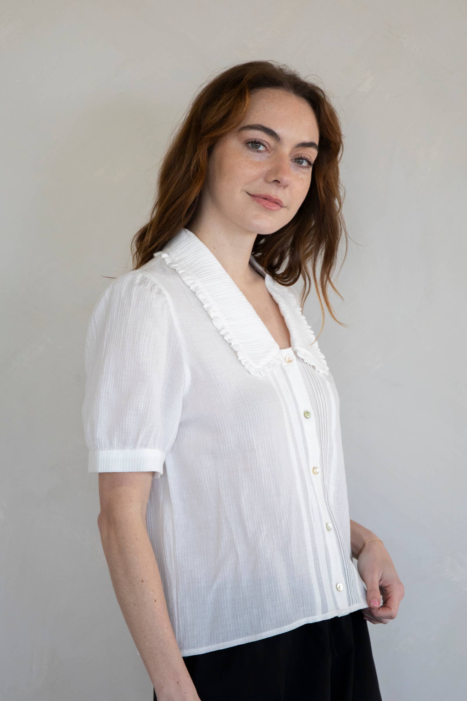Woman standing against white background wearing black shorts and white collared button down blouse with extended sleeves and vertical line details 