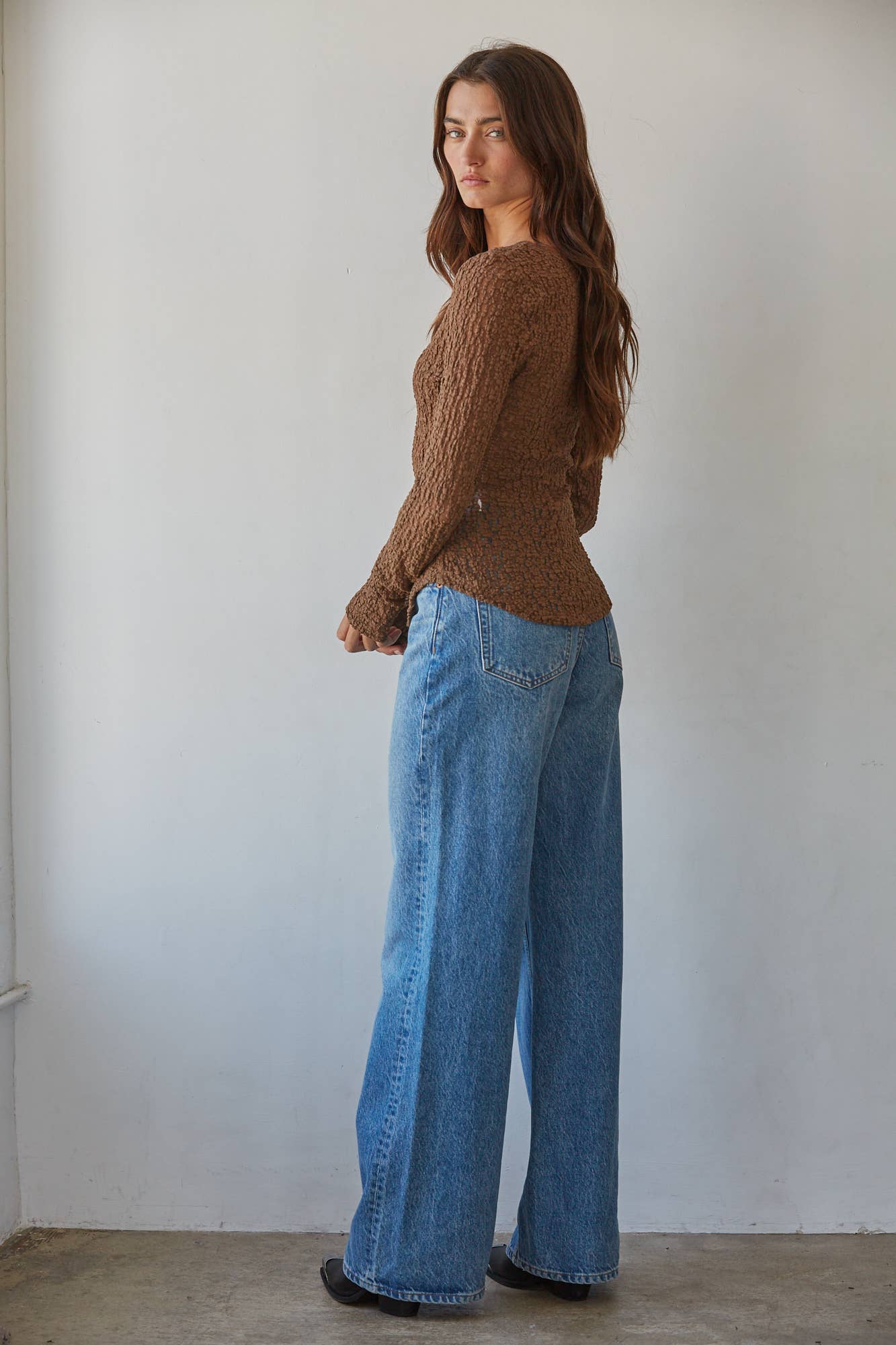 A woman stands in profile wearing a brown textured knit sweater and wide-leg blue jeans against a plain off-white wall.