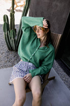 Woman wearing shorts and a dark Kelly green button up cardigan and blocking the Sun from her face 