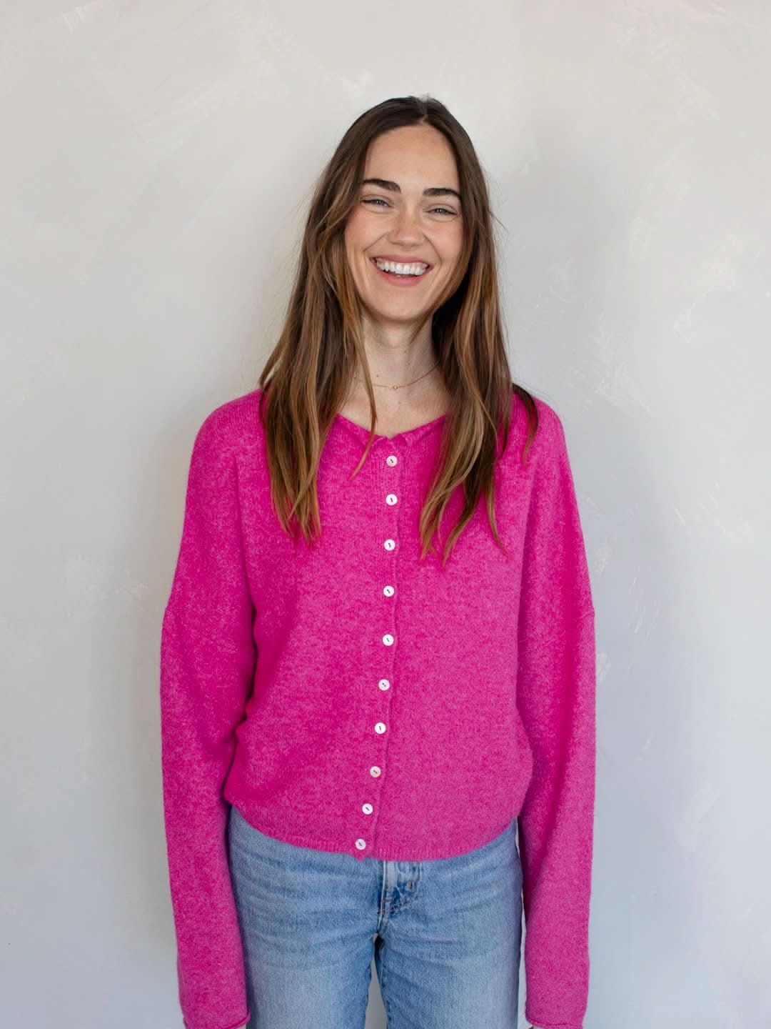 Woman smiling standing against white background wearing Fuschia pinkish button down cardigan with white buttons and blue jeans