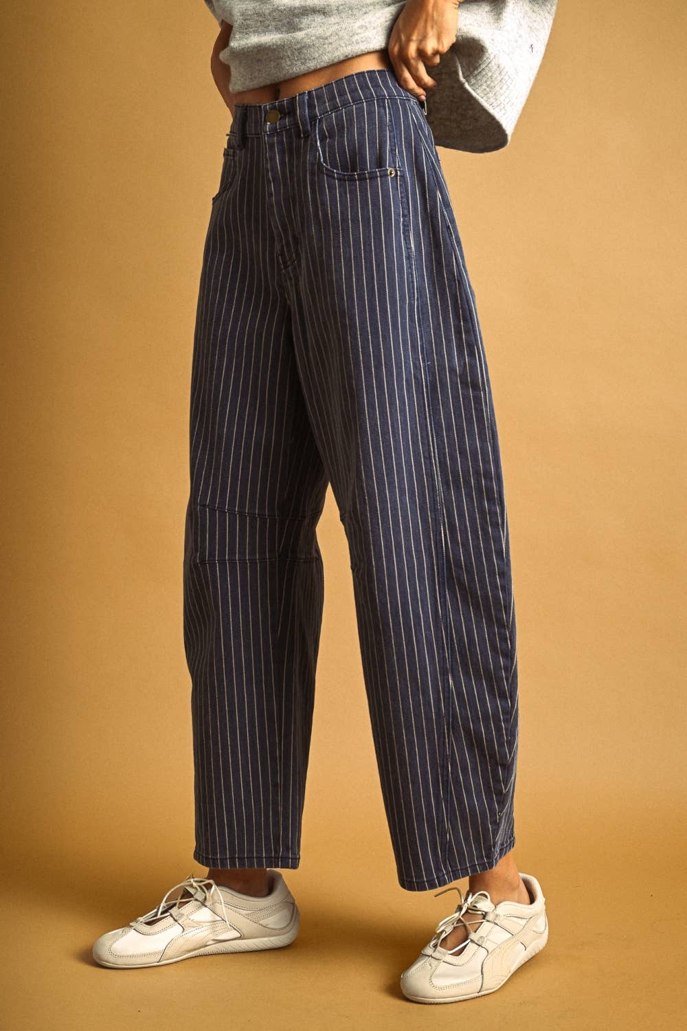 A person wearing navy blue pinstriped wide-leg jeans, a light gray knit top, and white sneakers, standing against a warm tan studio background.