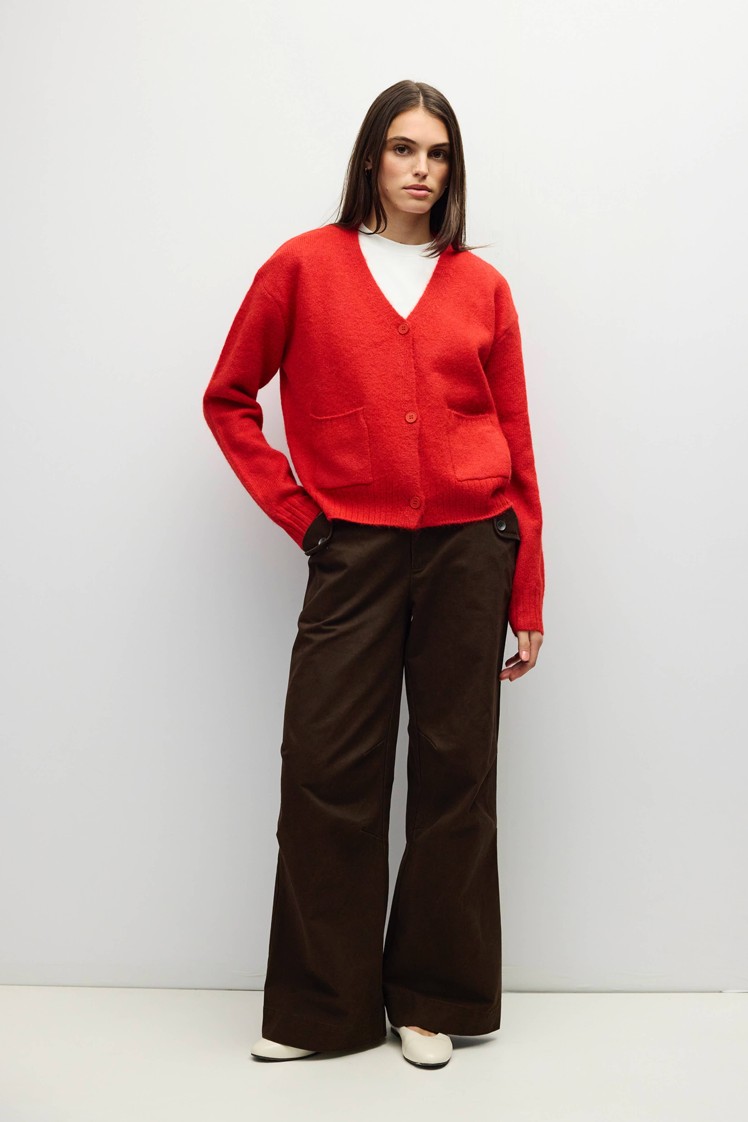 A woman stands against a light gray studio background wearing a bright red knit cardigan over a white top, paired with dark brown wide-leg trousers and white pointed shoes.