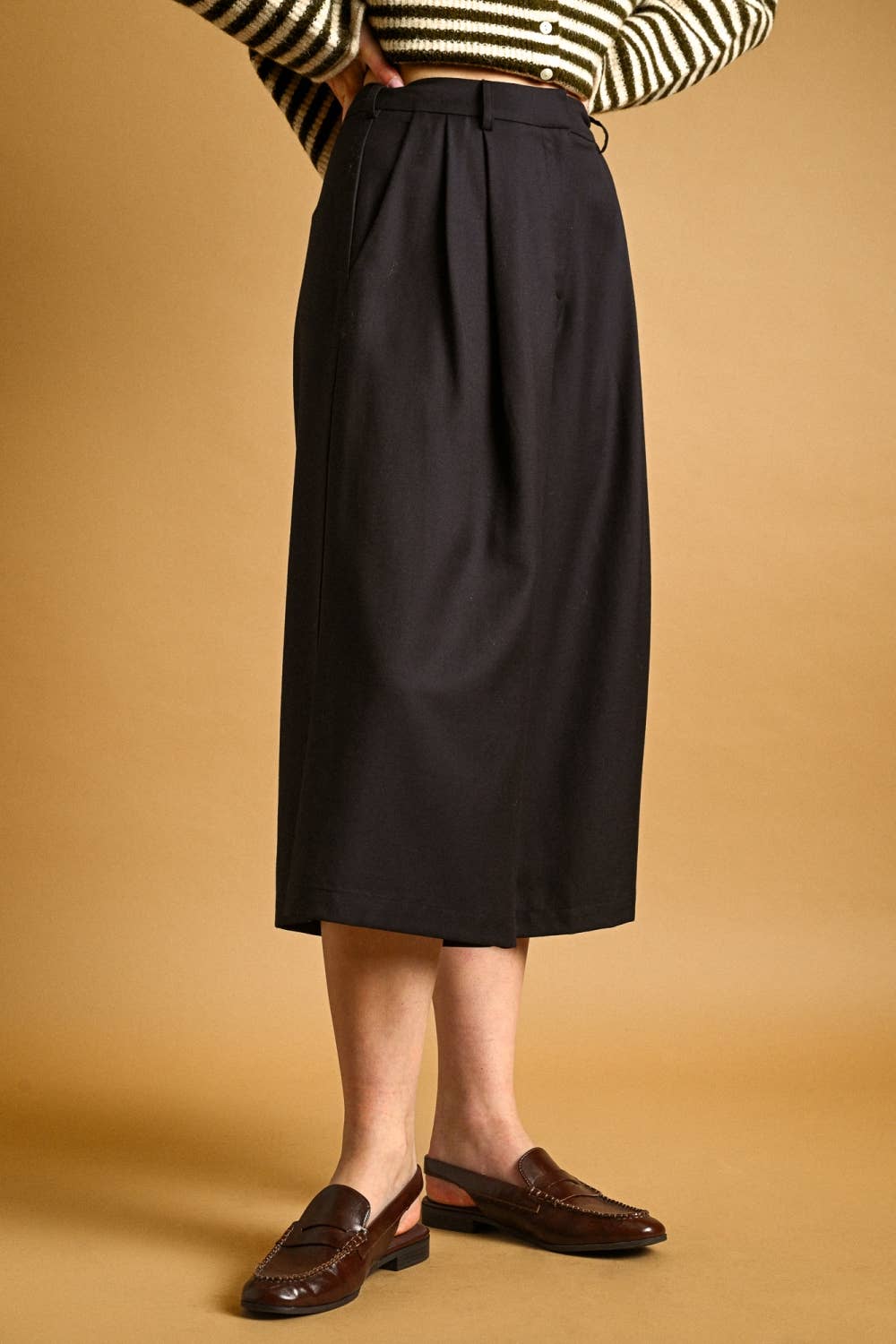 A person wearing a dark charcoal pleated midi skirt, a black and white striped sweater, and brown leather loafers, standing against a solid tan background.