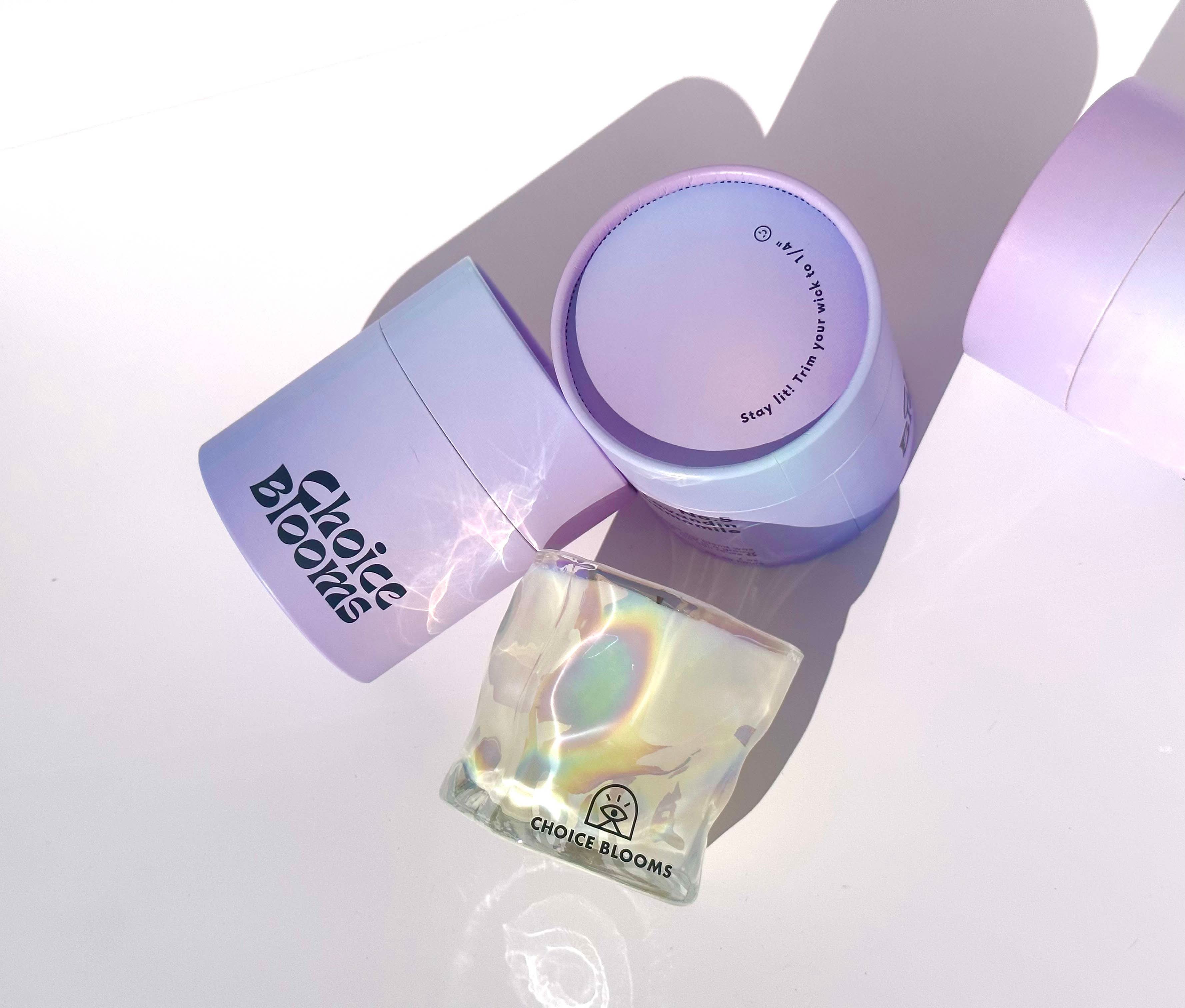 One candle in iridescent wavy glass cup laying on its side next to two purple packaging containers 