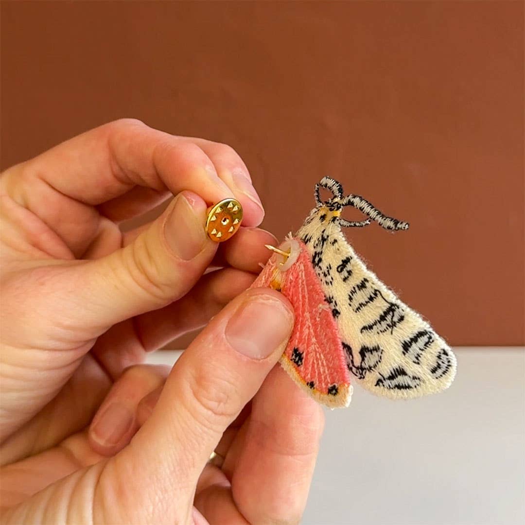 Hand holding a small embroidered moth with pink wings and black patterns against a brown background