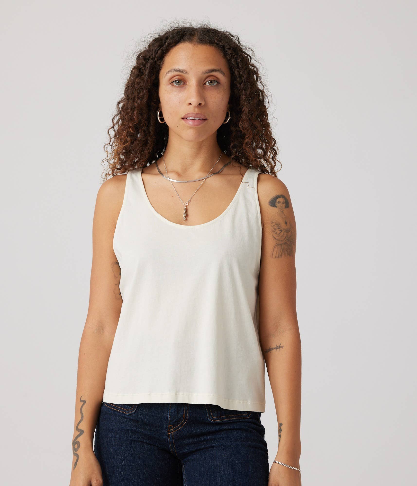 Woman wearing a white tank top and dark jeans against a plain background