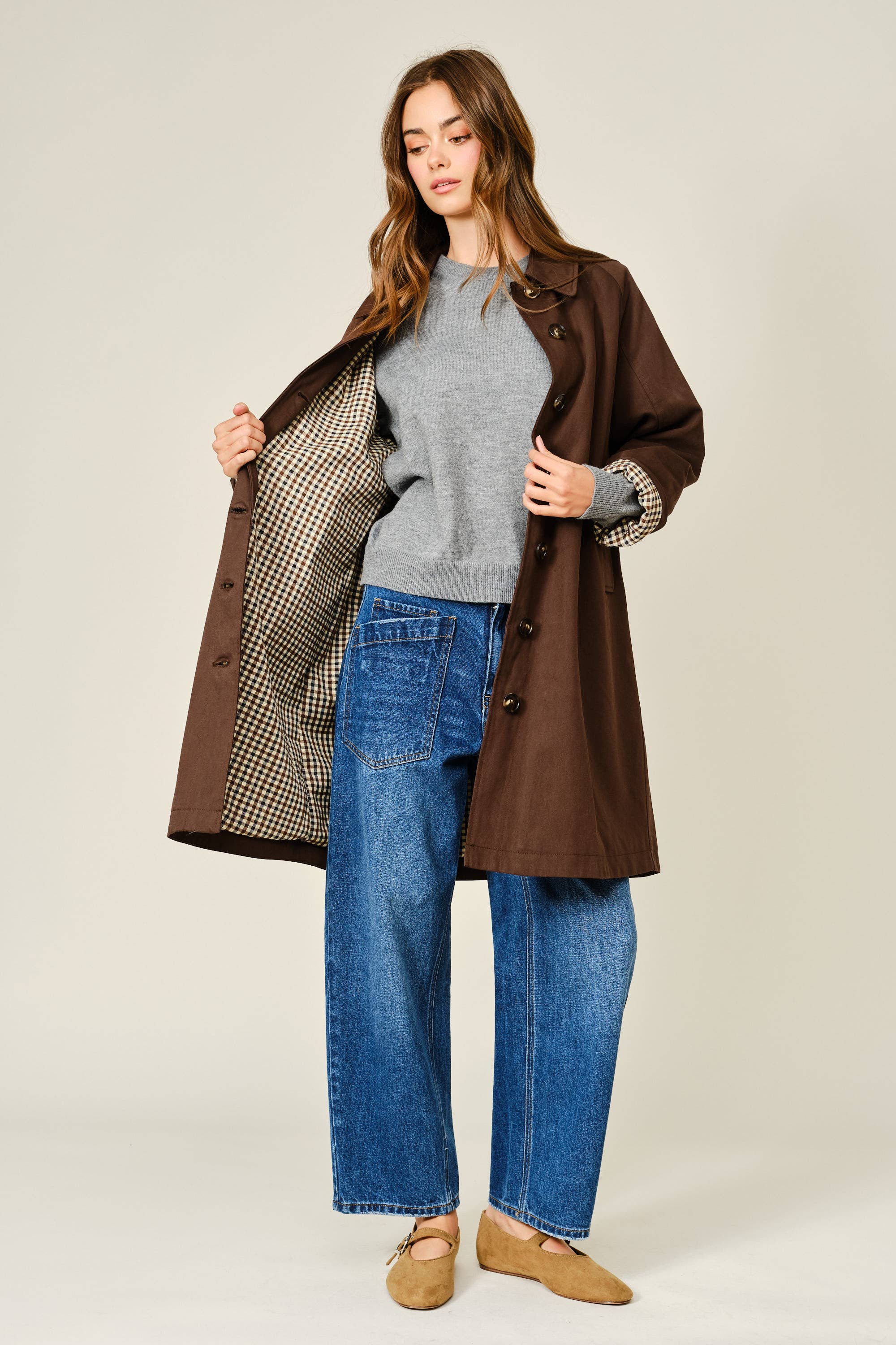 Woman wearing a brown coat over a gray sweater and blue jeans on a white background