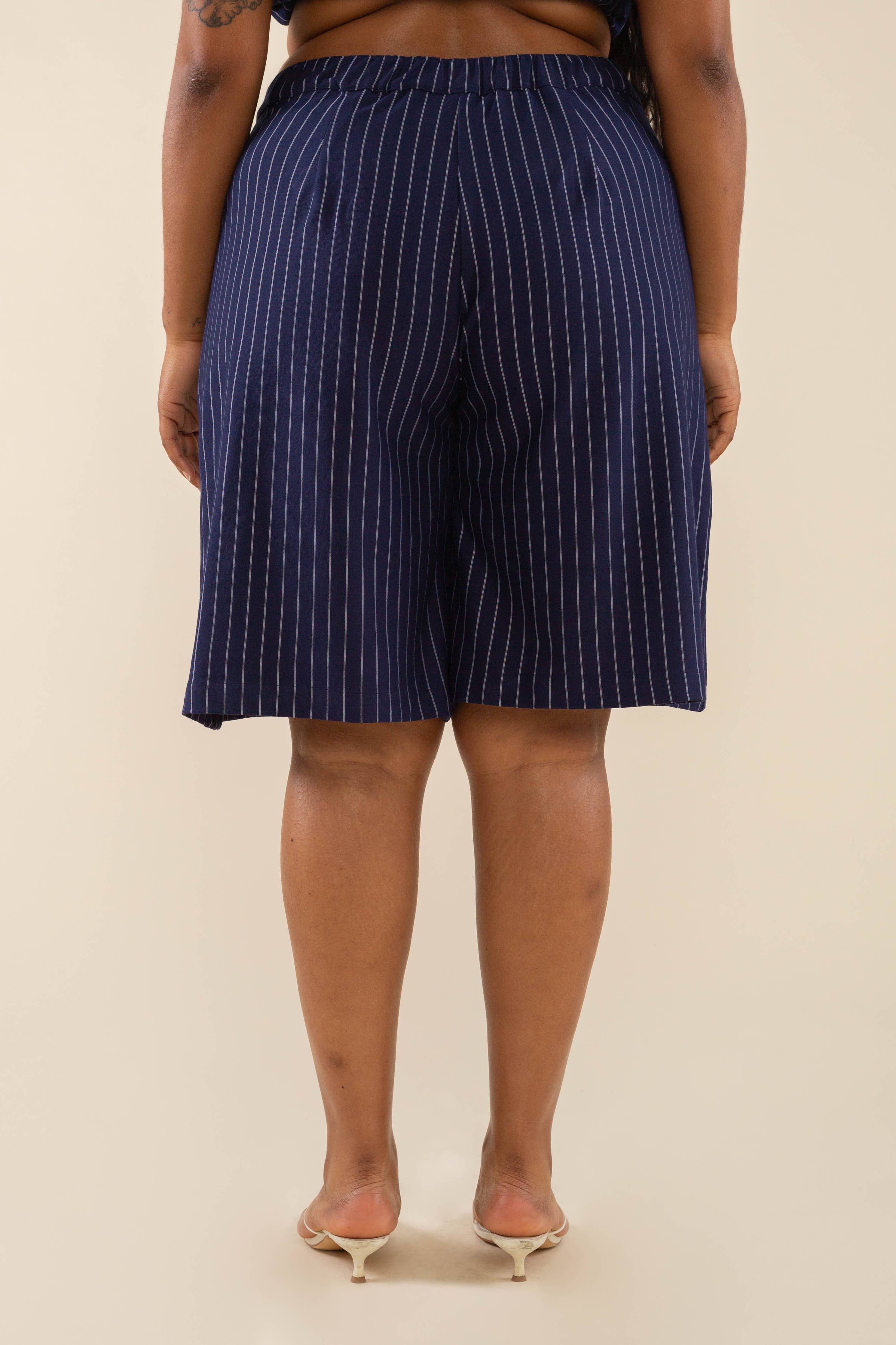 Woman wearing navy vertical pin striped knee length shorts and white heels behind a blank background. 