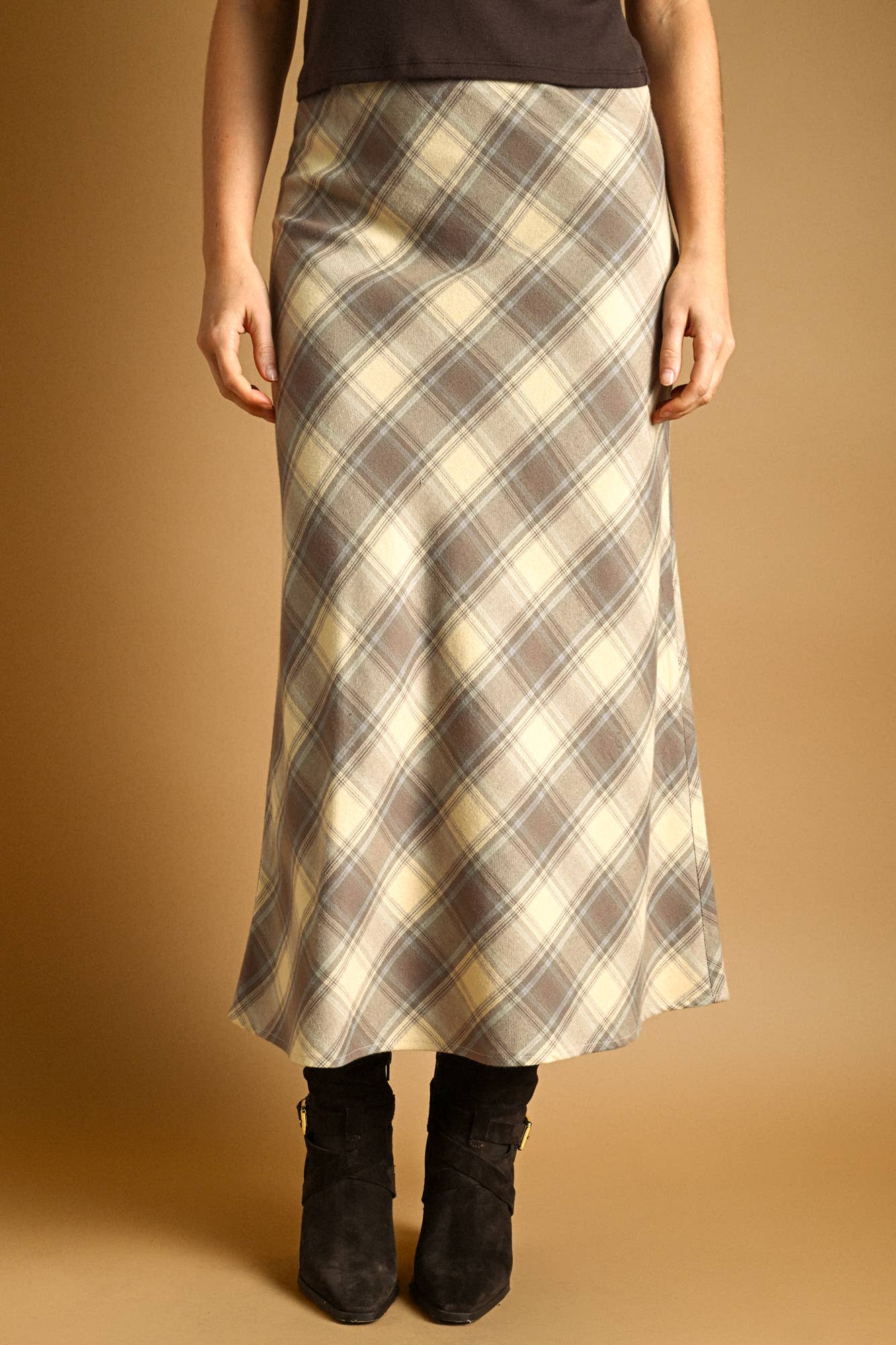 A woman wearing a long plaid midi skirt in beige, brown, and cream tones, paired with black ankle boots, against a solid warm brown studio background.