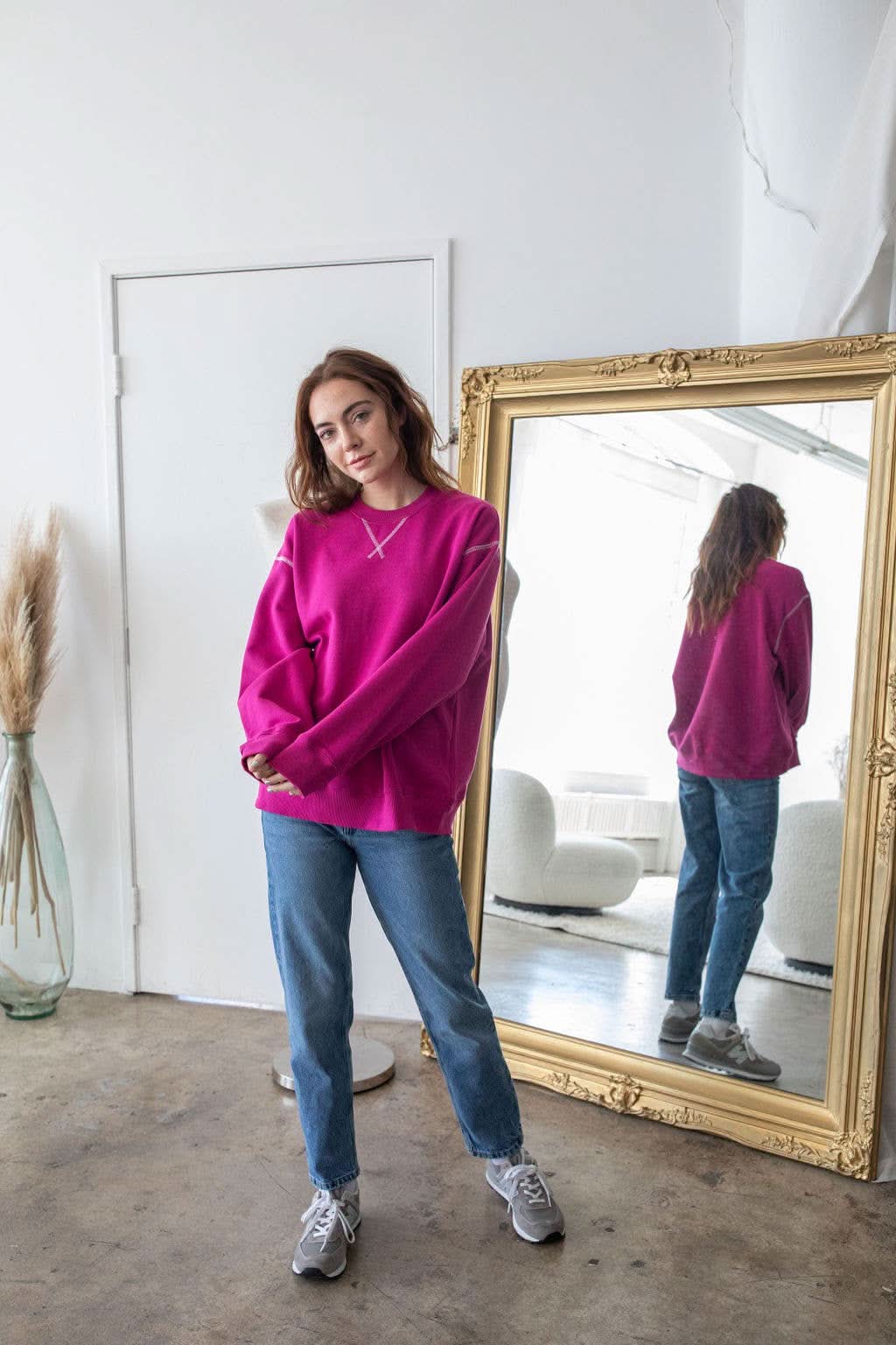Woman wearing pinkish purple pullover with unique stitching details standing next to a gold framed mirror 