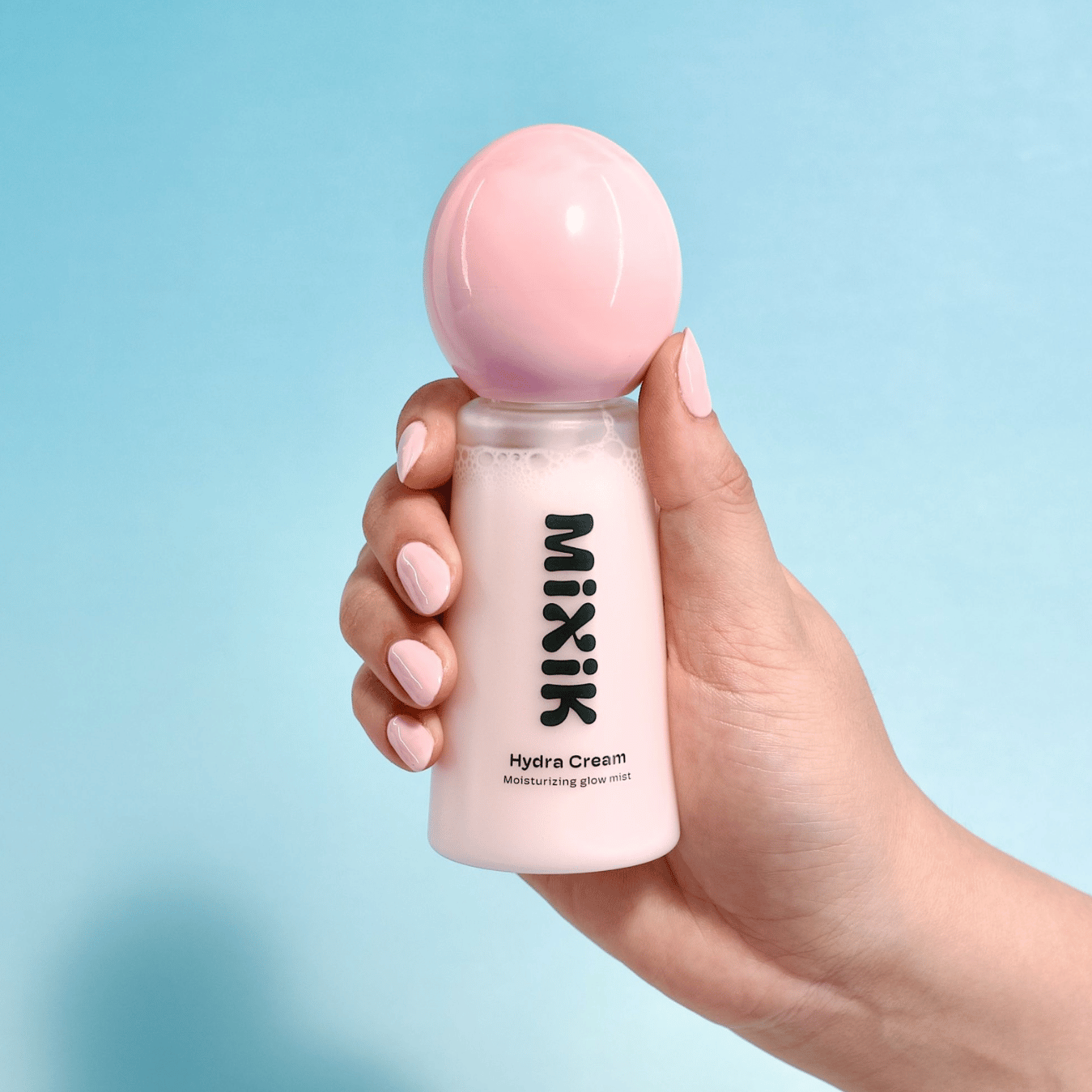 Hand holding a pink bottle of Mixik Hydra Cream against a light blue background