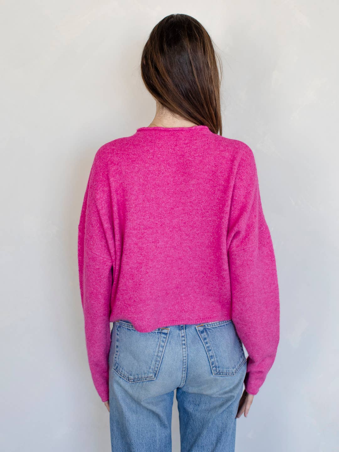 Woman smiling standing against white background wearing Fuschia pinkish button down cardigan with white buttons and blue jeans