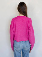 Woman smiling standing against white background wearing Fuschia pinkish button down cardigan with white buttons and blue jeans