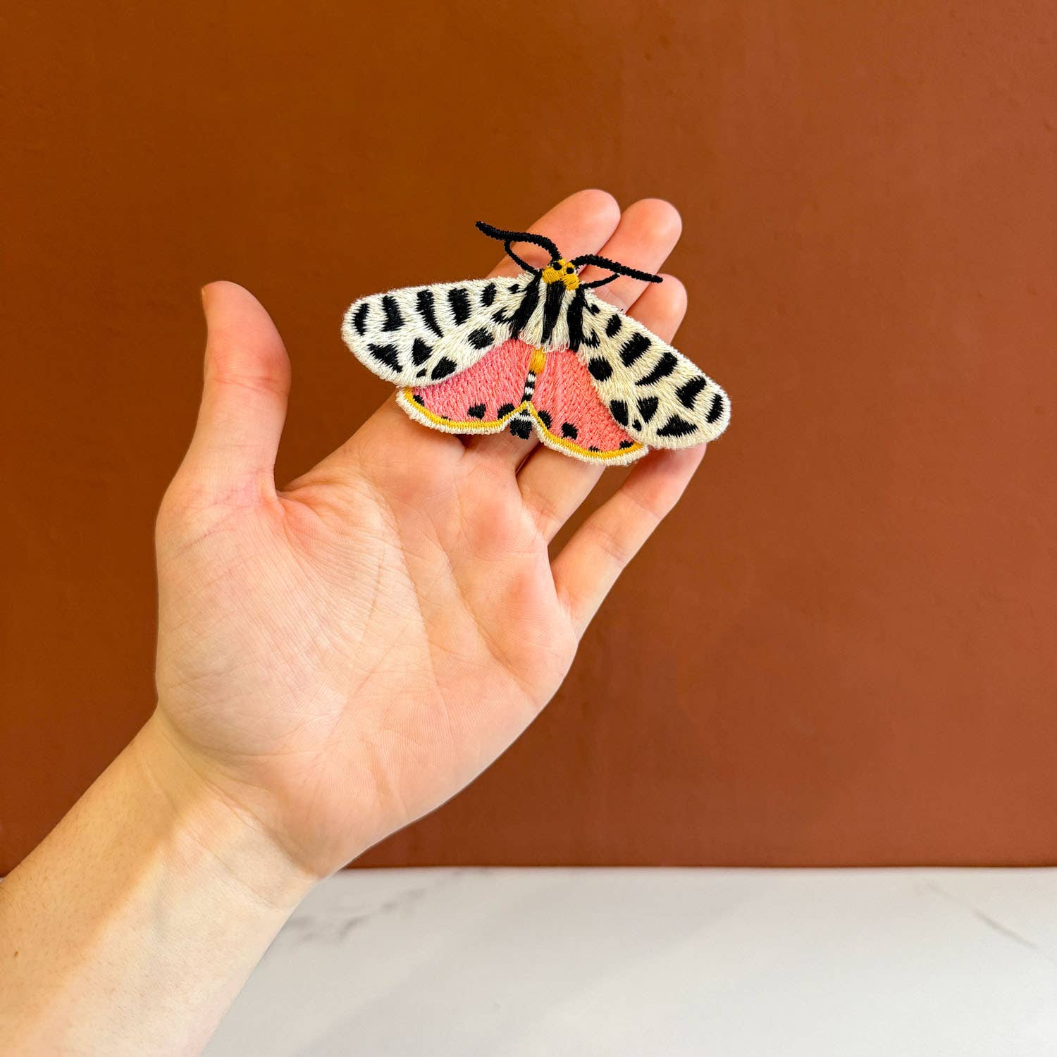 Hand holding a colorful moth-shaped patch against a brown background