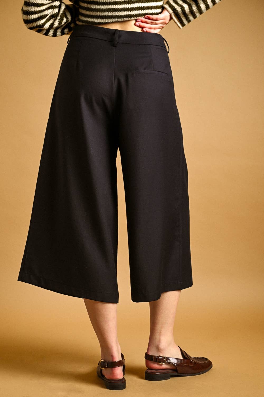 A person wearing black wide-leg cropped pants with a striped top and brown sandals, standing against a warm tan studio background.