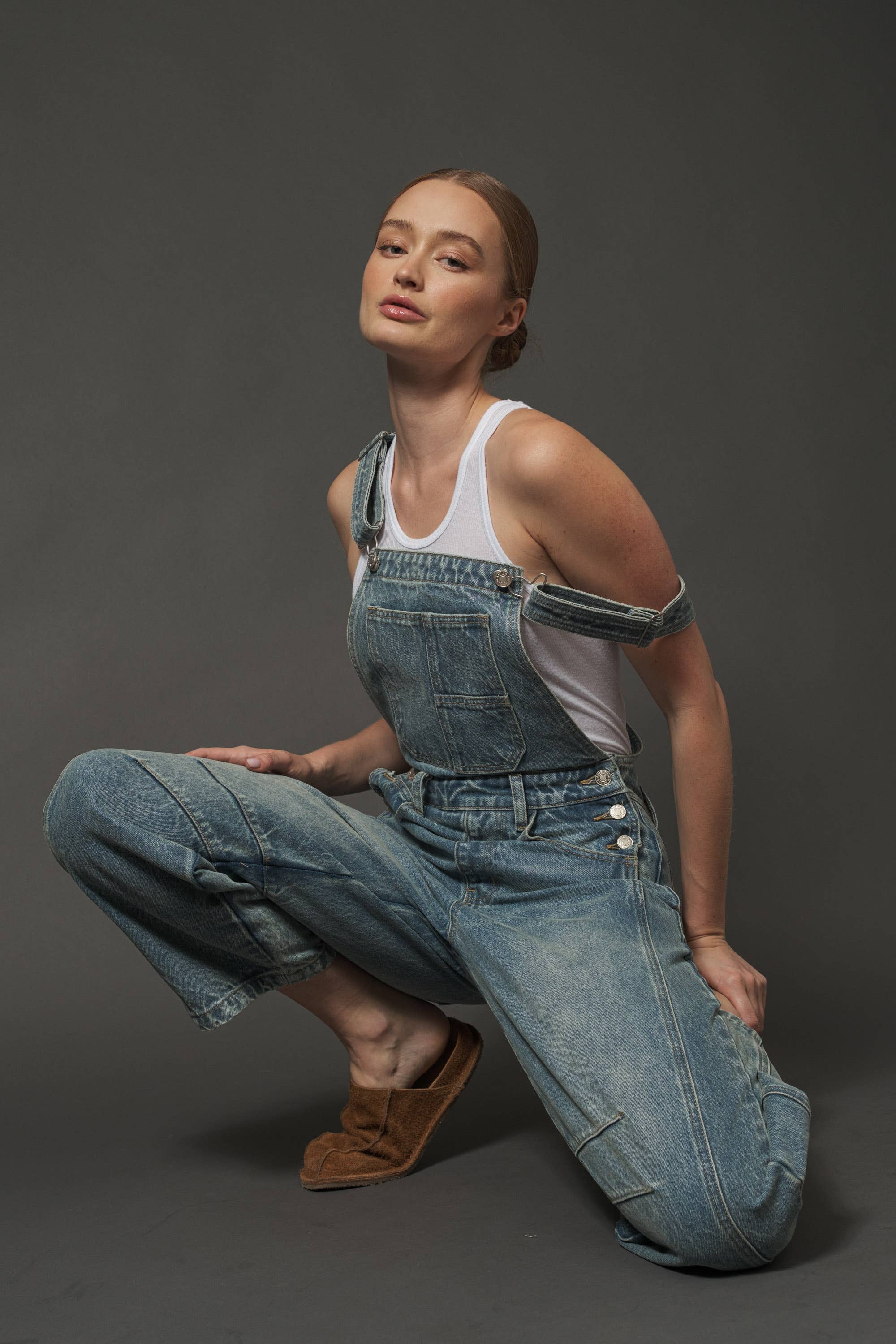 Person wearing blue denim overalls and a white tank top on a dark background