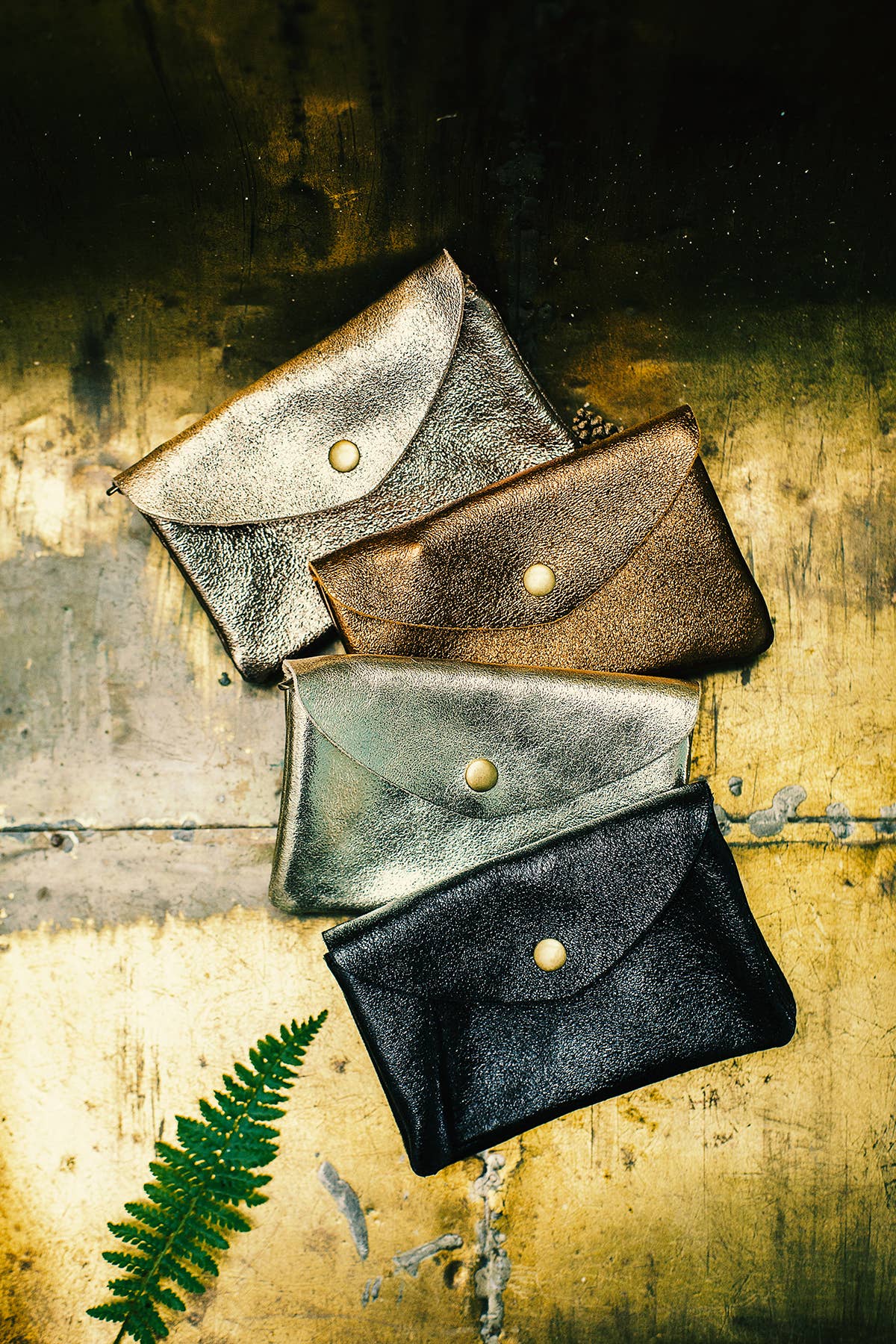 Four small leather envelope-style pouches or card cases in metallic silver, bronze, gray, and black, arranged diagonally on a distressed gold surface with a green fern leaf at the lower left.
