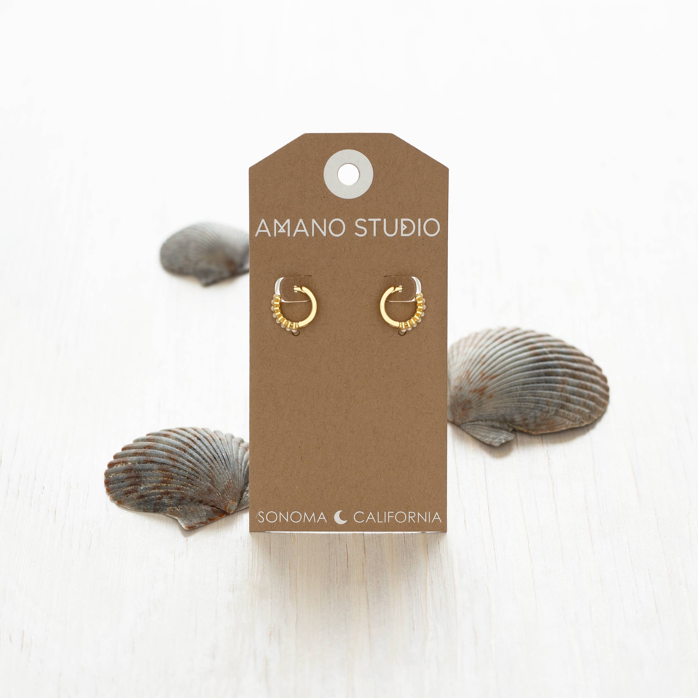 Gold hoop earrings on a 'Amano Studio' branded card with seashells in the background.