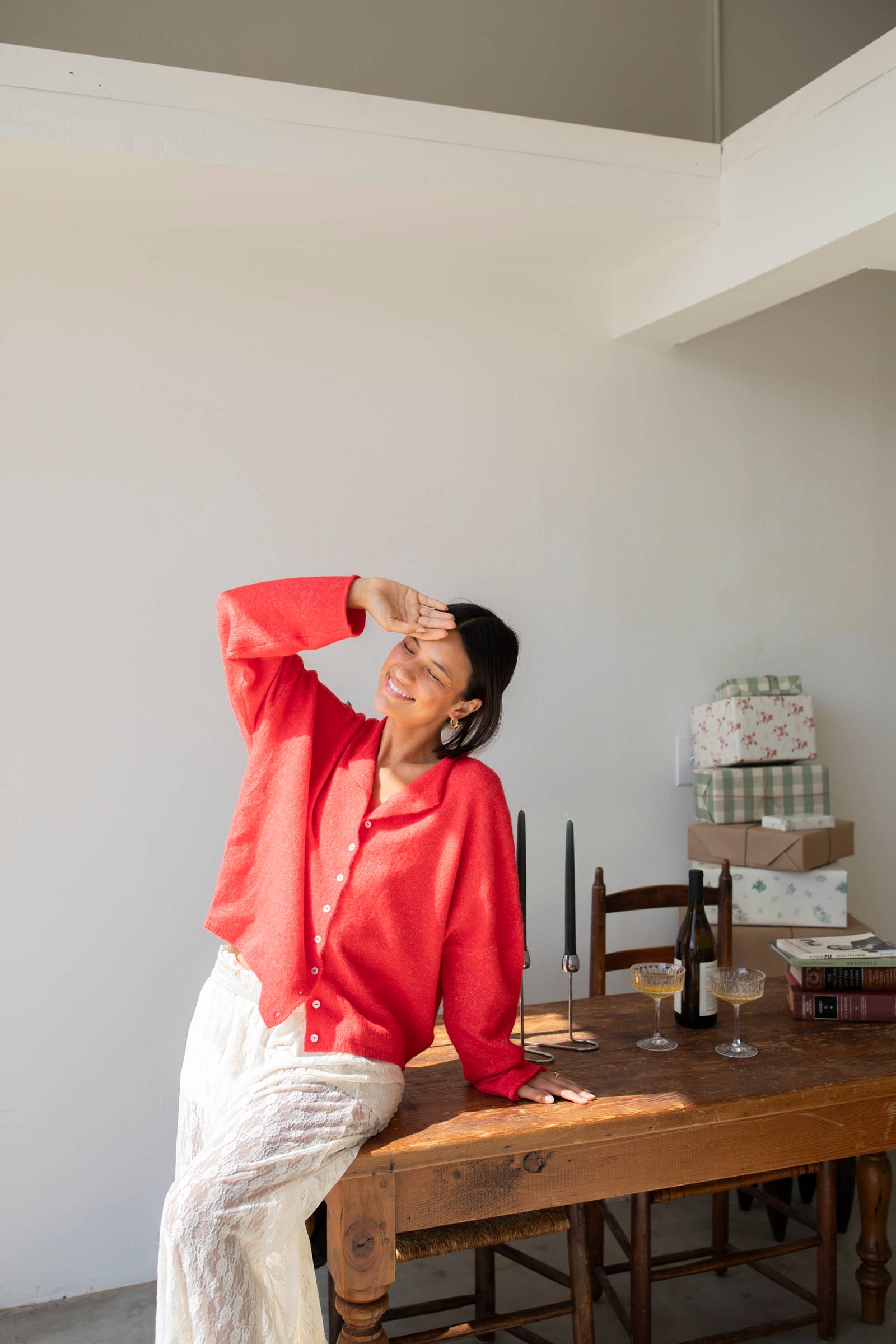 Woman smiling standing against white background leaning on a wood table wearing red button down cardigan with white buttons and white mesh skirt 