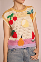 Woman wearing sleeveless, ivory, colorful, crochet, top with cherries, lemon, strawberry, watermelon, pear, and pineapple with pink, purple, and yellow horizontal stripes 