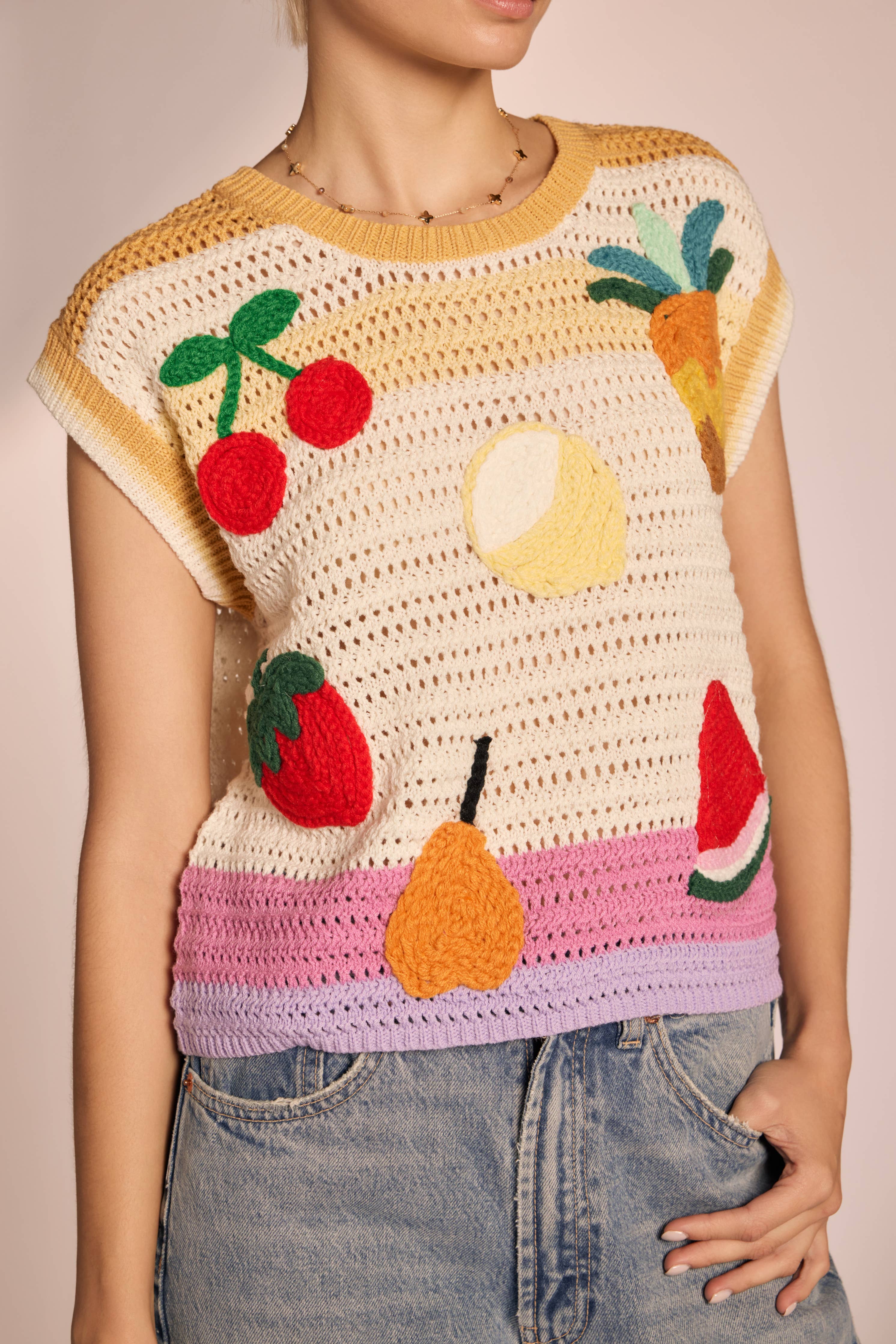 Woman wearing sleeveless, ivory, colorful, crochet, top with cherries, lemon, strawberry, watermelon, pear, and pineapple with pink, purple, and yellow horizontal stripes 
