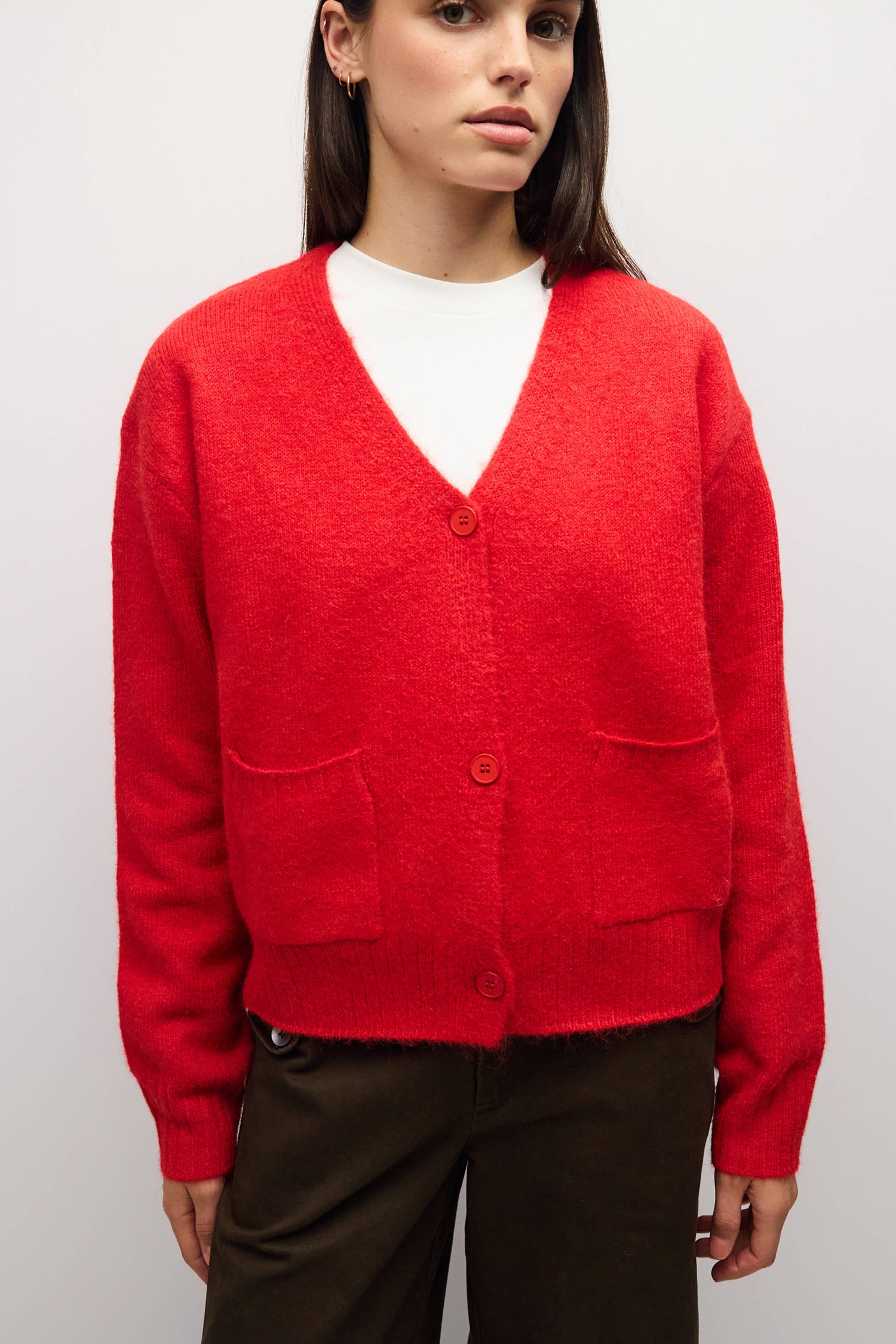 A woman wearing a bright red fuzzy knit cardigan with a V-neck, three buttons, and front patch pockets, layered over a white top and dark brown pants, photographed against a light gray studio background.