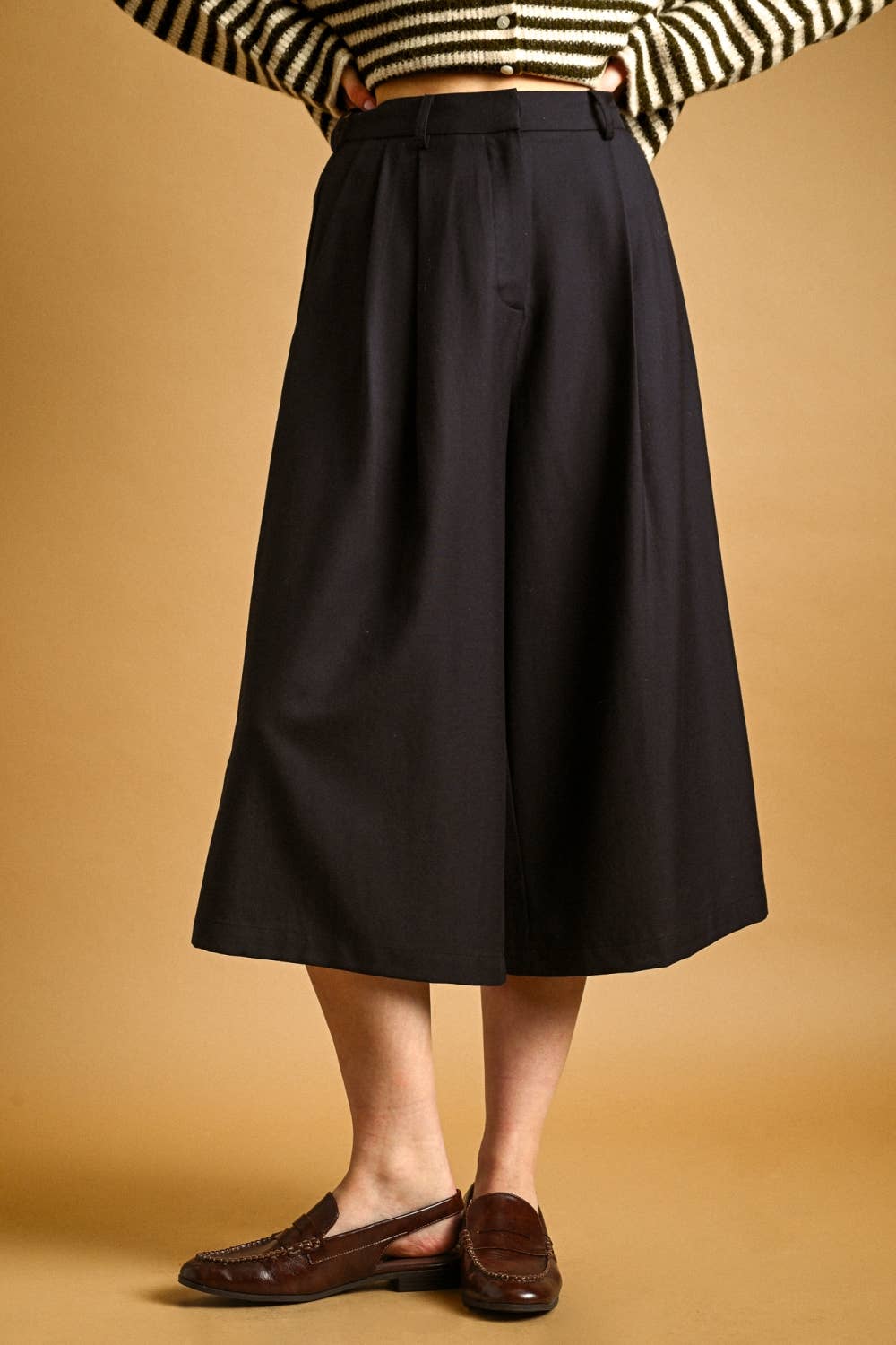 A person wearing a black wide-leg midi skirt, styled with a black and white striped top and brown leather loafers, against a solid warm tan background.