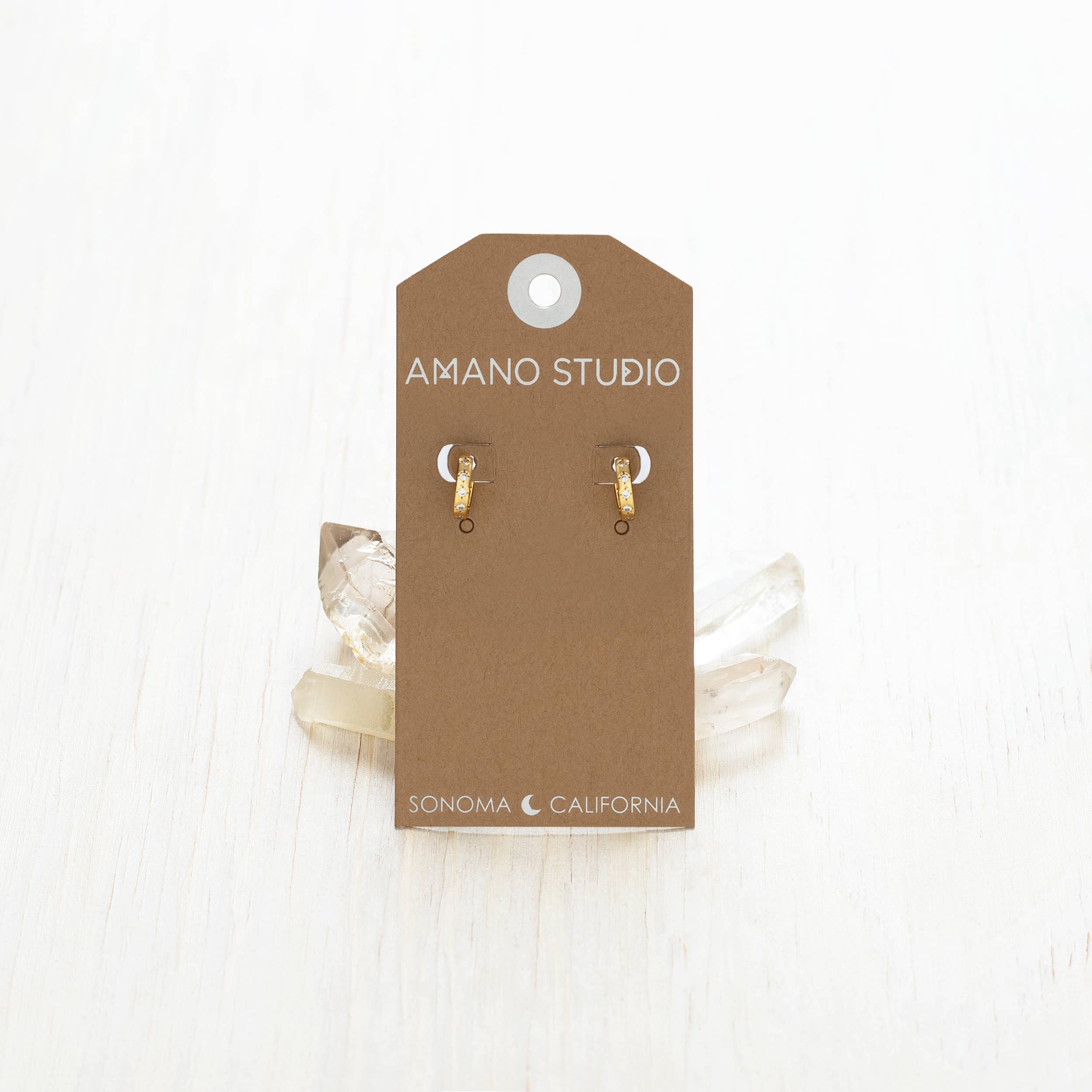 Gold earrings on a brown Amano Studio card with white lace in the background