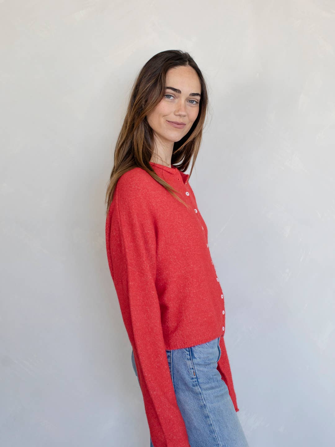 Woman smiling standing against white background wearing red button down cardigan with white buttons and blue jeans
