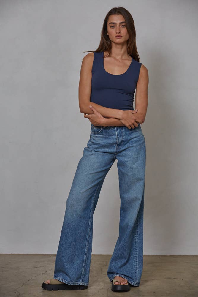 Woman wearing a blue tank top and jeans standing against a plain background
