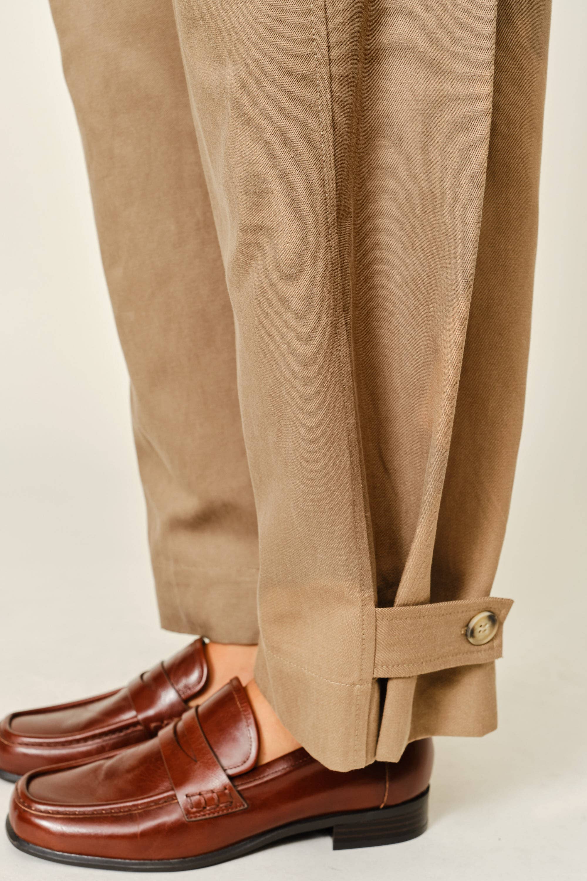 Close-up of a person wearing tan trousers with a buttoned cuff and brown leather penny loafers, shown against a plain light beige background.